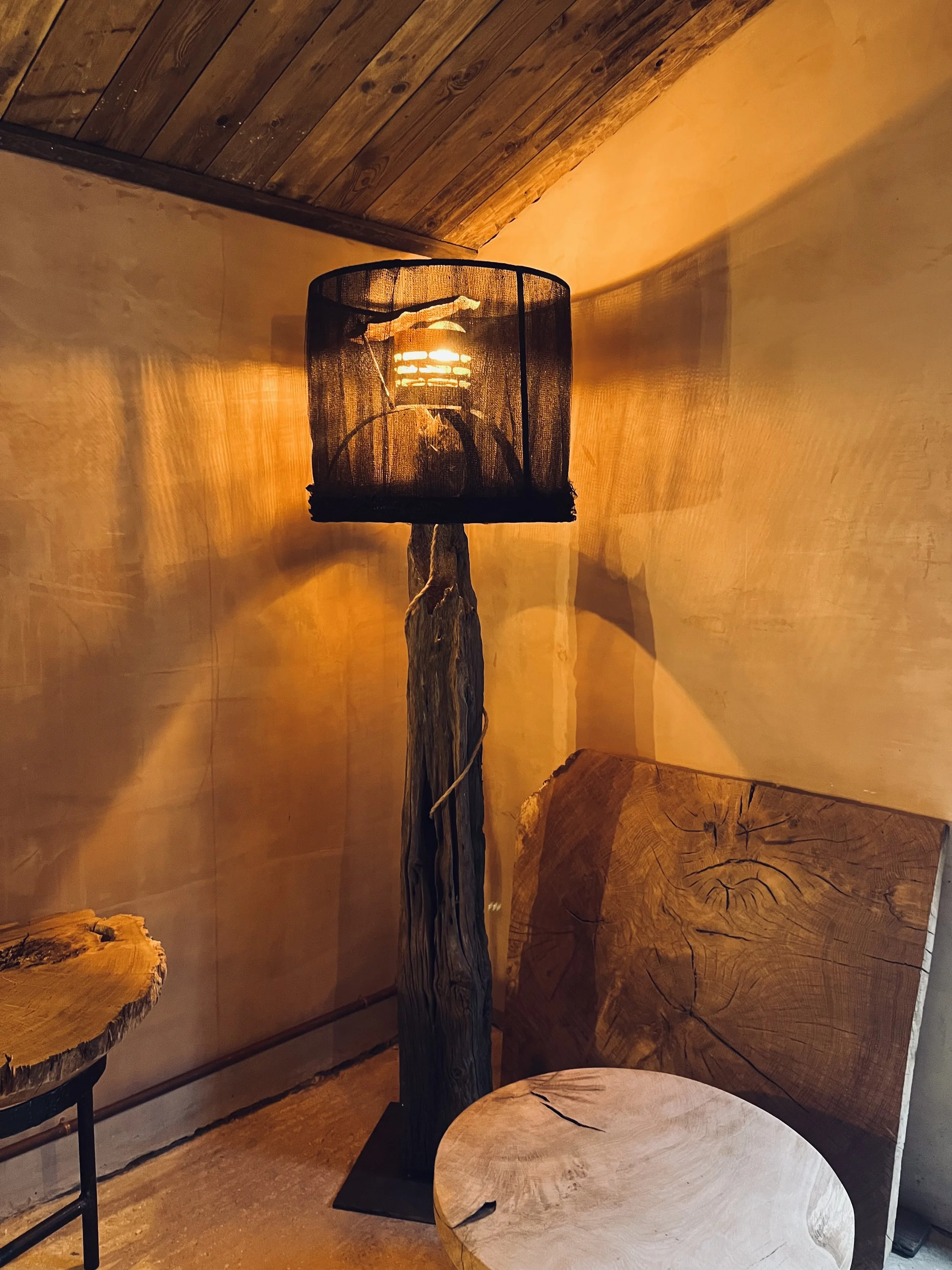 A rustic floor lamp made from a twisted tree trunk with a fabric shade encasing the light bulb, placed in a cozy room with wooden furniture and warm lighting.