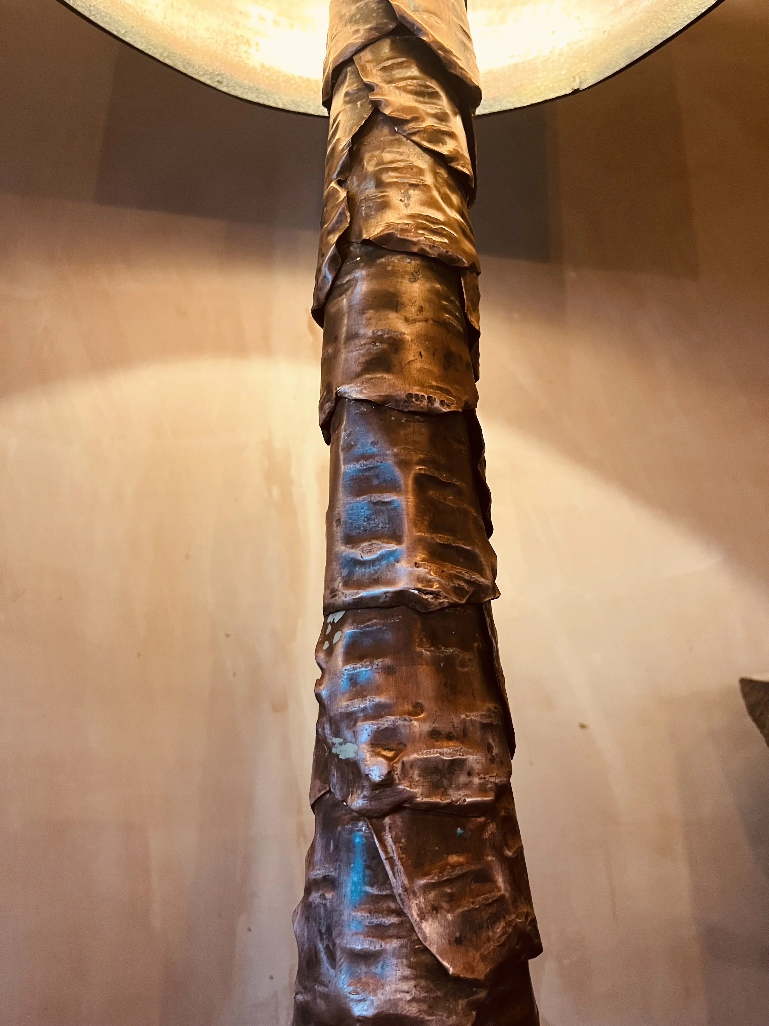 Close-up of a copper lamp post with a textured, bark like design, twisted surface and a warm glow at the top.