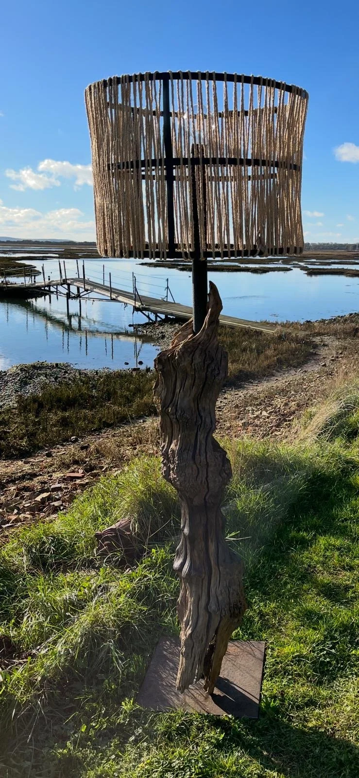 A wooden lamp with a rustic, twisted wooden base and a cylindrical lampshade made of vertical wooden slats, standing outdoors near a body of water on a sunny day.