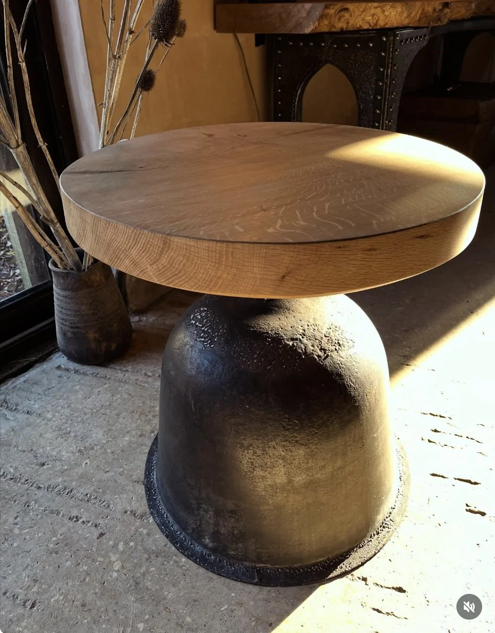 A round wooden table with a textured base, placed on a concrete floor near a window with sunlight shining on it.