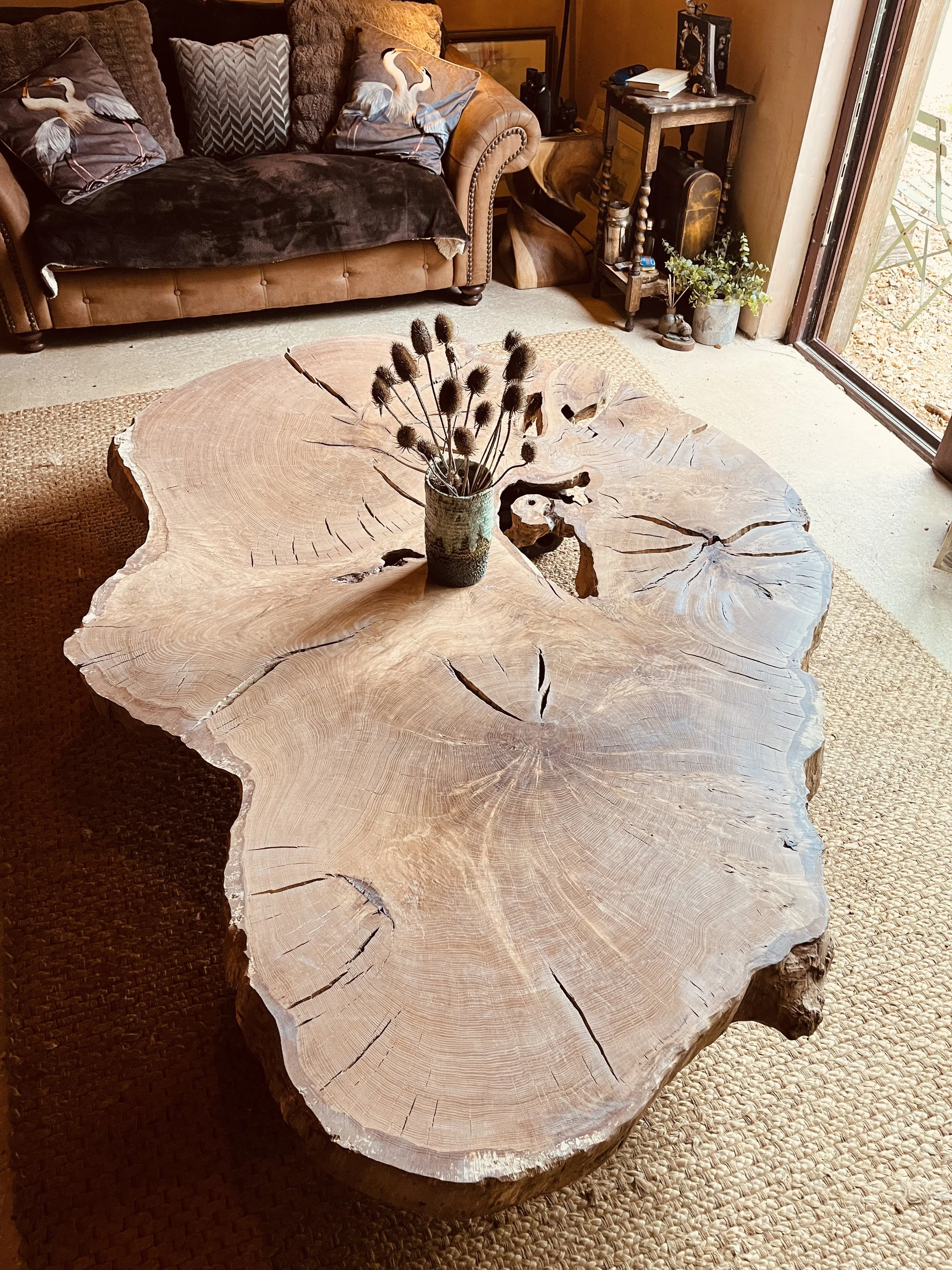A large wooden coffee table with a natural, uneven edge and visible growth rings, decorated with a jar of dried thistle flowers, is in a cozy living room. In the background, there is a brown leather sofa with decorative pillows, a small side table with plants and books, and a sliding glass door leading outside.