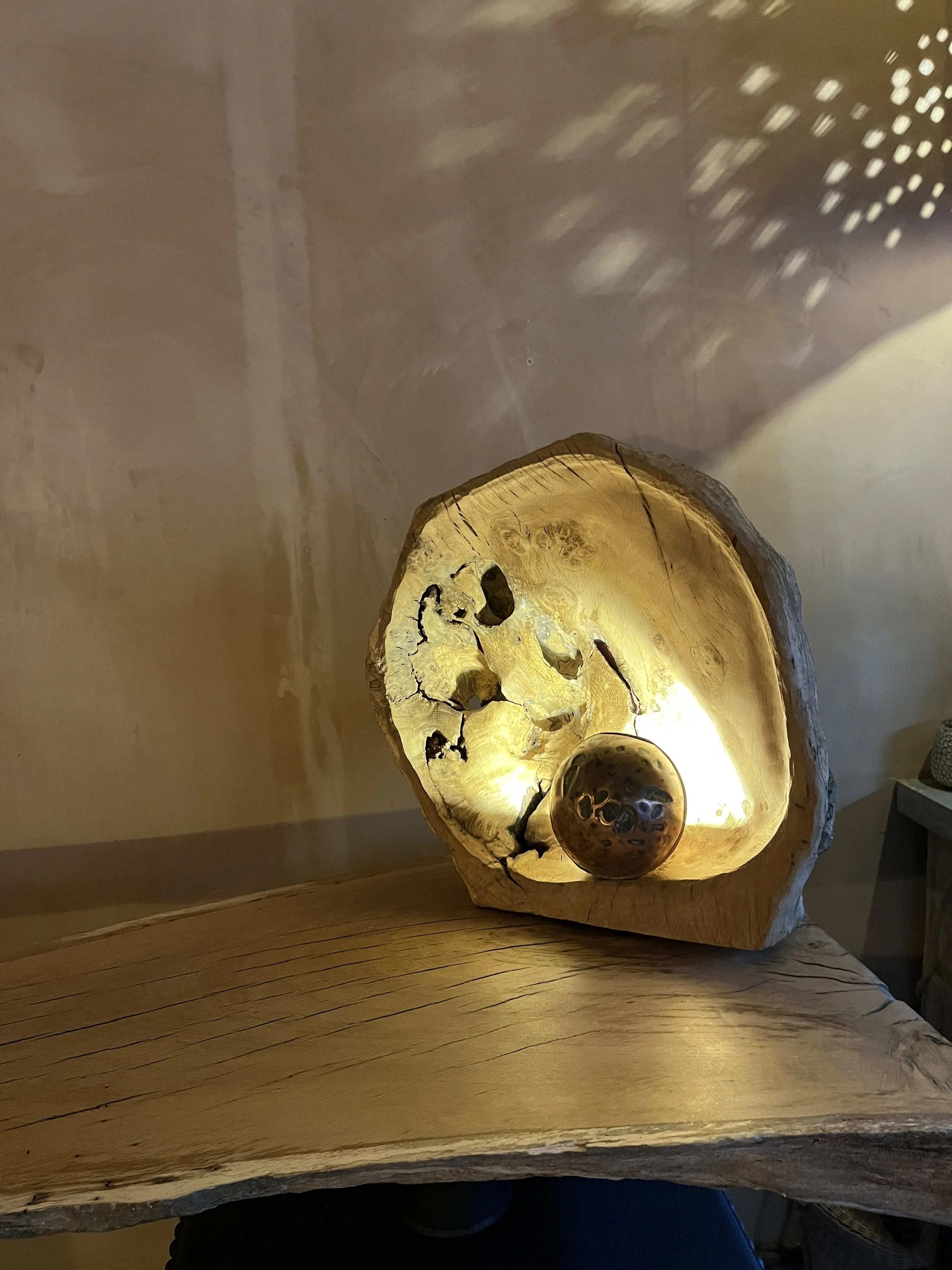 A decorative lamp made from a large piece of wood with natural cracks and a hollowed-out center, illuminated from within, with a small metallic sphere resting inside the hollow.