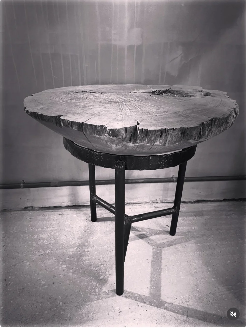 A rustic wooden table with a thick, rough edge and three black metal legs, placed against a grey wall with an industrial aesthetic.