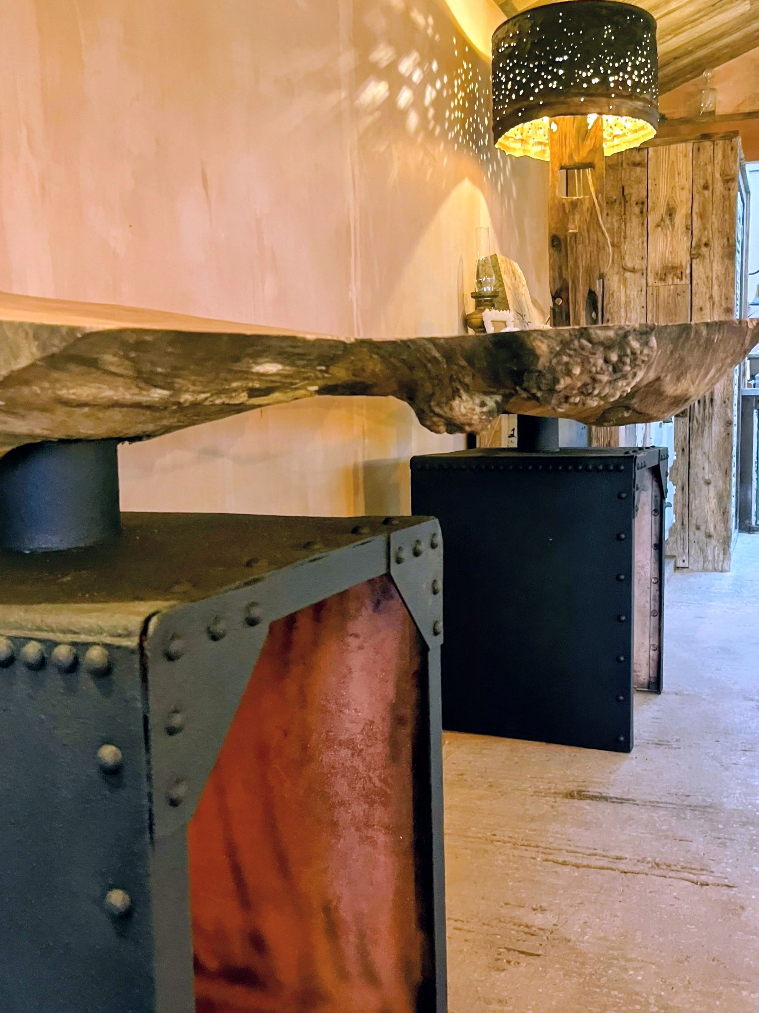 Close-up of a rustic wooden table with a thick, uneven edge, supported by black metal bases with rivets. In the background, there is a decorative lamp with a perforated black shade casting patterned light on the wall, and a wooden cabinet.