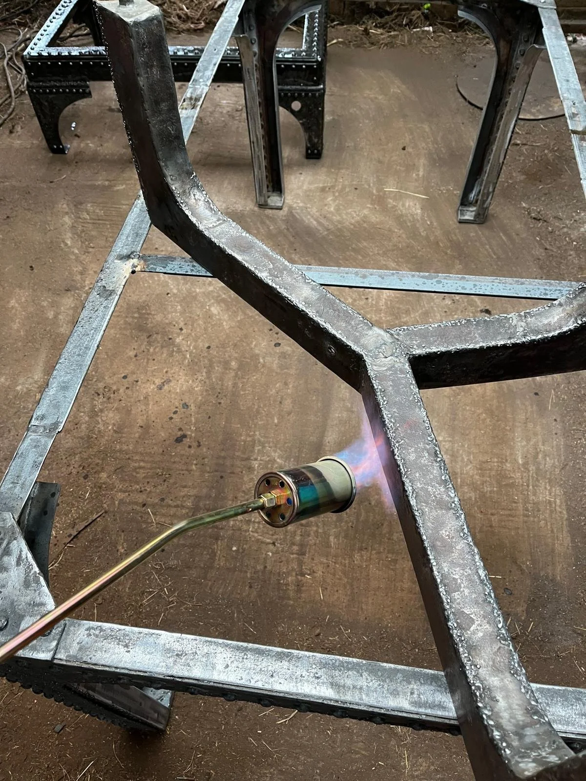 Welding a metal frame with a handheld torch, sparks and heat visible, in a workshop with a dirt floor.