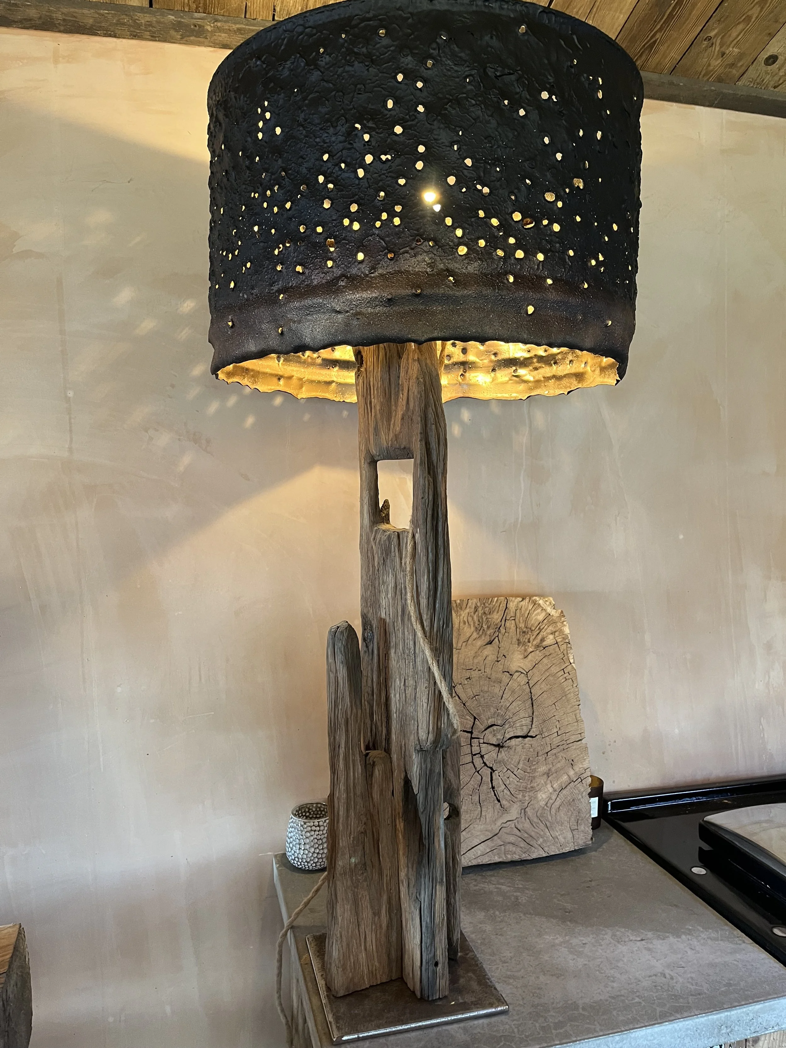 A lamp with a black perforated lampshade and a wooden base made of driftwood on a gray surface.