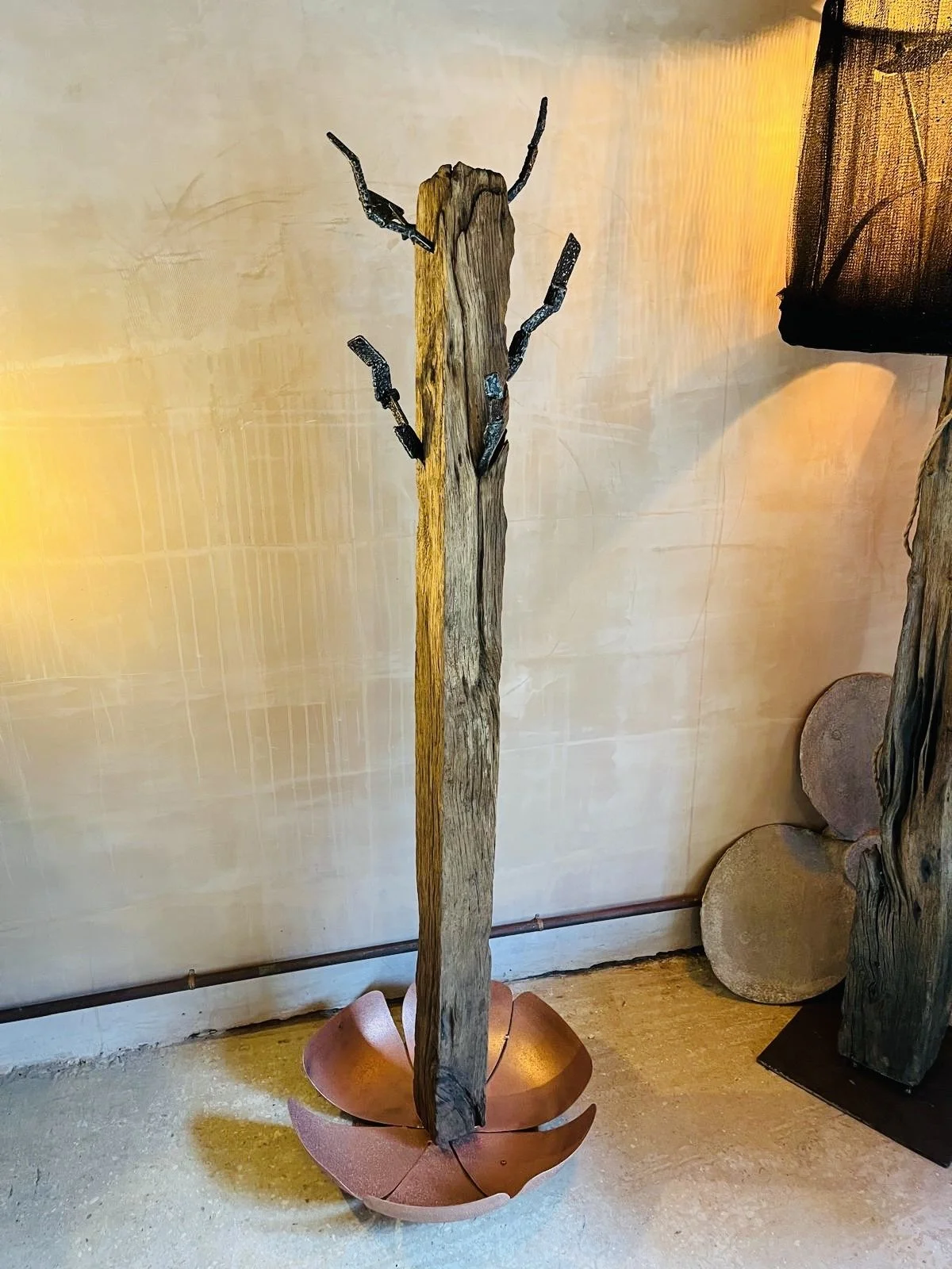 Decorative sculpture of black metallic knives embedded in a tall broken piece of weathered wood, mounted on a curved, reddish-brown base resembling an abstract flower or shell, inside a room with a textured beige wall and minimal lighting.