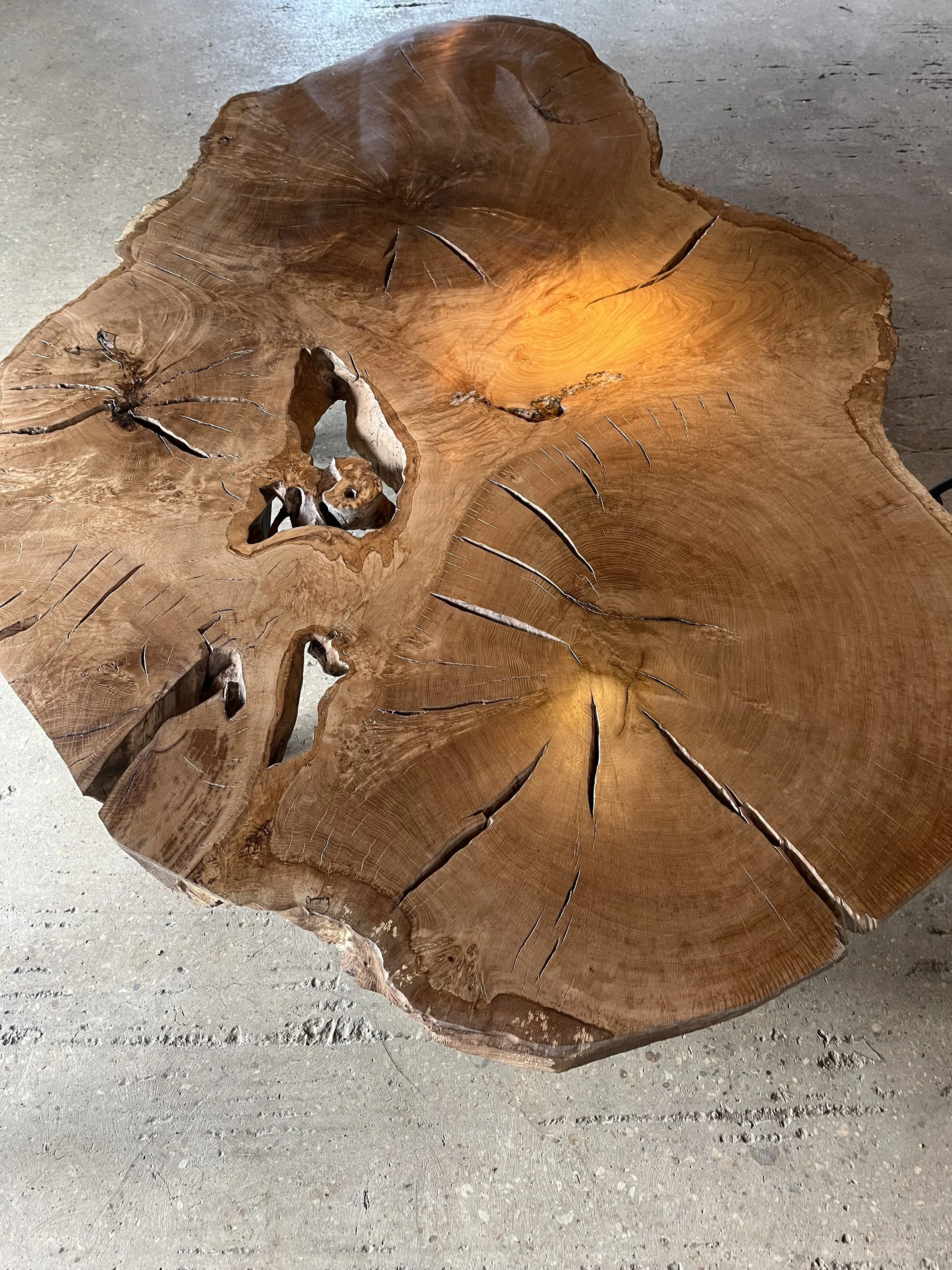 A large round wooden table with natural cracks and holes, showcasing the wood grain and rings, placed on a concrete floor.