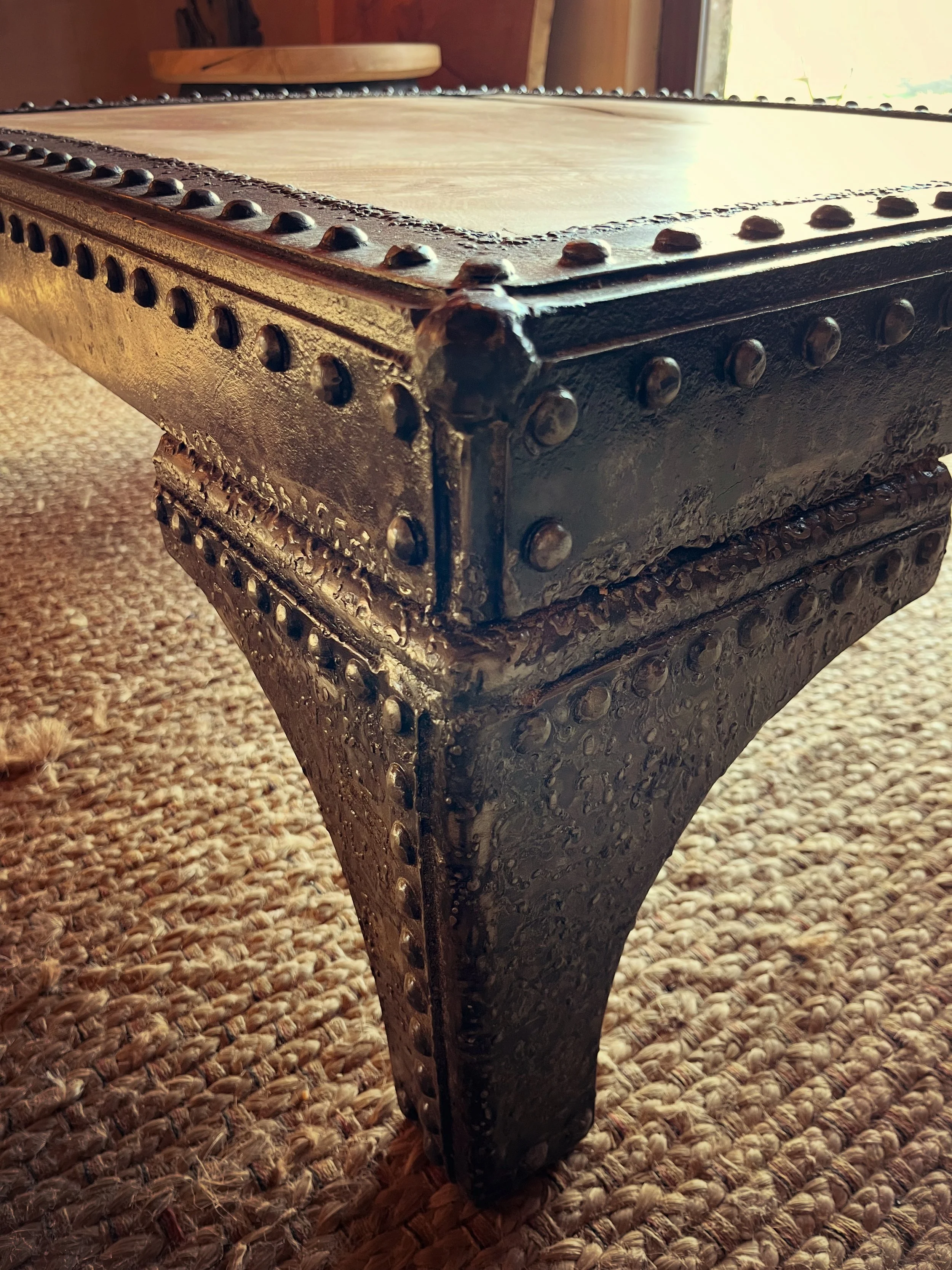 Close-up of a metal table or bench with rivets, showing corner and leg, placed on a woven carpet, with a room and window in the background.