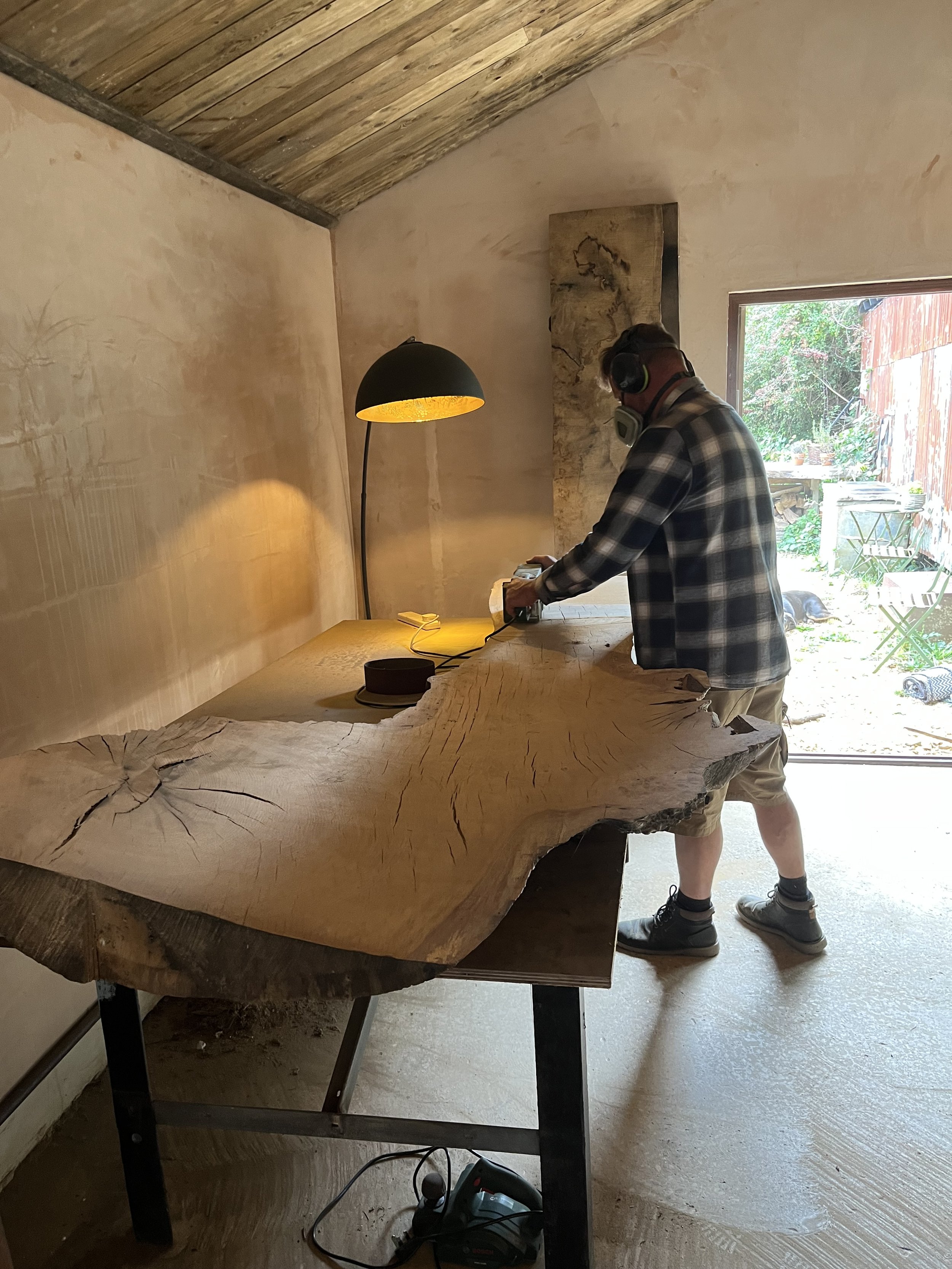 A man wearing headphones and a plaid shirt uses a power tool on a large, natural wood slab table in a woodworking shop or studio, with a floor lamp illuminating the workspace.