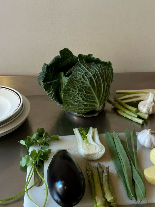 Groene groenten en kruiden, waaronder een kool, lente-uitjes, knoflook, groene bonen, asperges, waterkers, en een aubergine op een kookplaat en snijplank. Veel vezels en gezonde voeding