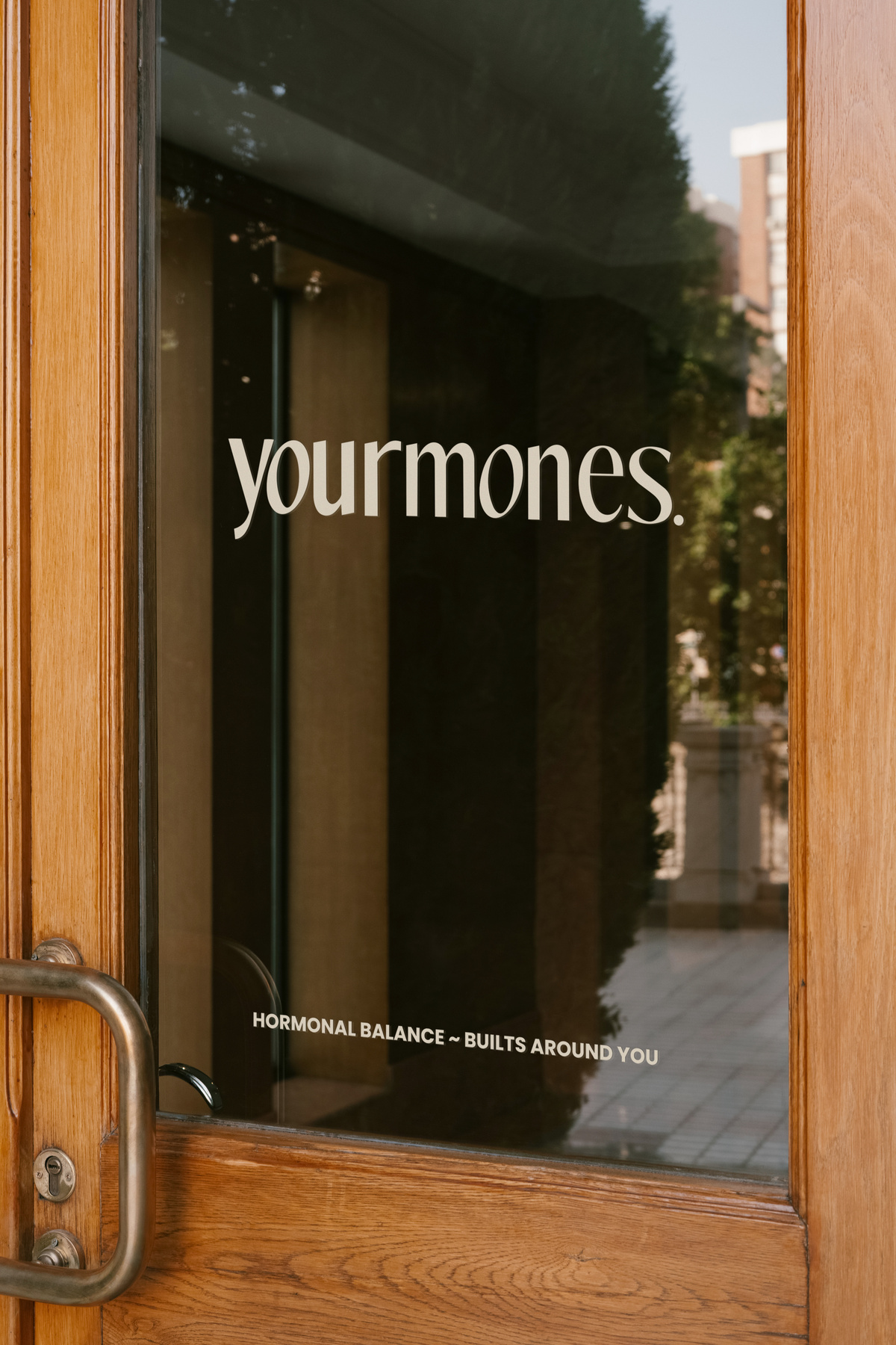 Deur met glas en houten frame met de woorden 'yourmones.' in wit letters en onderaan de tekst 'HORMONAL BALANCE ~ BUILT AROUND YOU'.