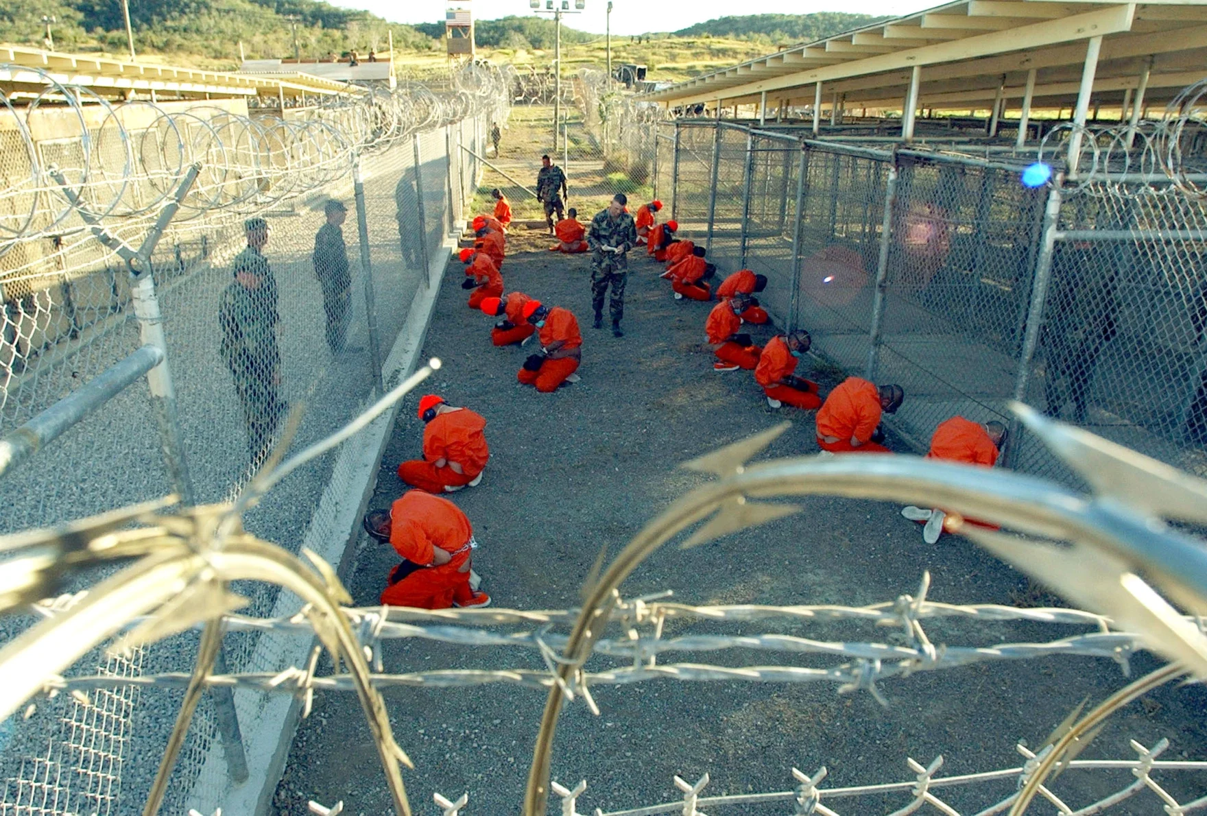 Prisoners dressed in orange jumpsuits kneeling and bowing their heads inside a fenced compound within a detention facility, with guards standing nearby.
