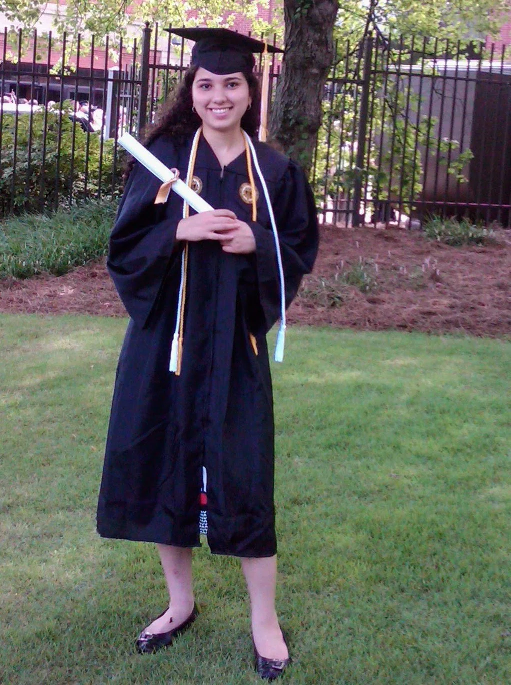 A young woman in black graduation gown and cap holding a diploma, standing on grass with trees and a black fence in the background.