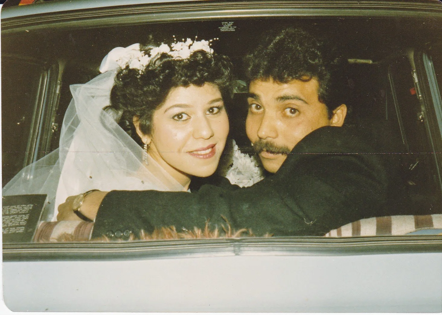 A bride and groom sitting in a car, celebrating their wedding day, with the bride smiling and wearing a veil and floral headpiece, and the groom looking at the camera.