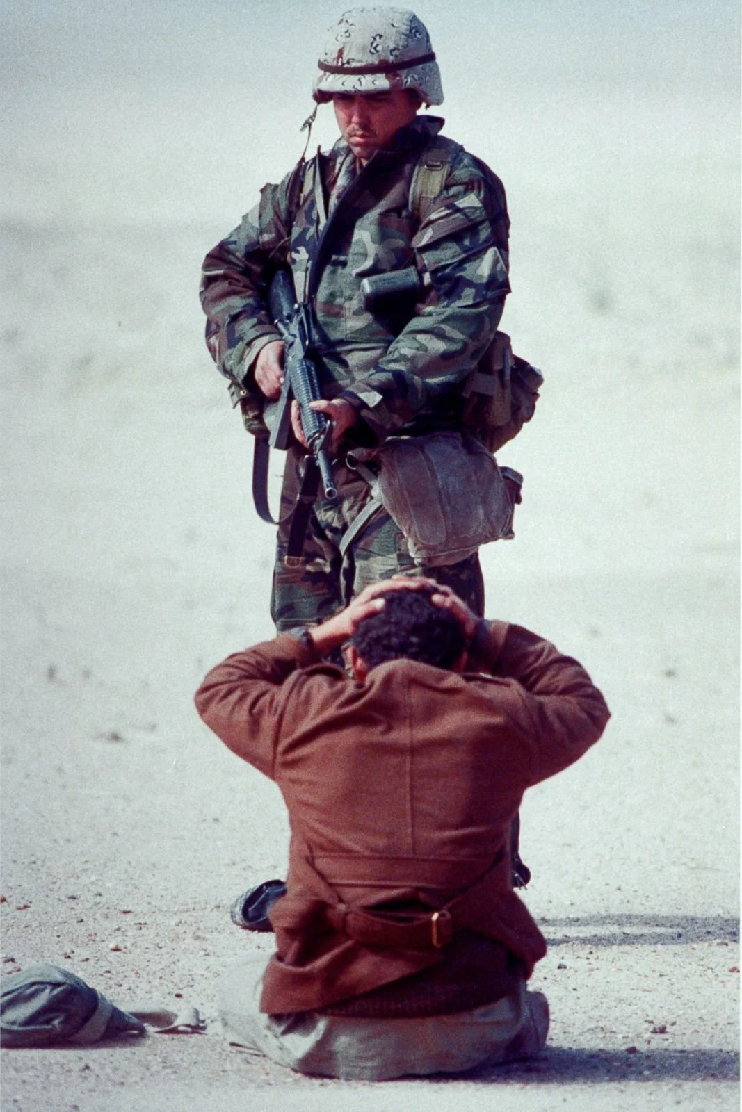 A soldier in camouflage uniform and helmet standing with a rifle, looking down at a person kneeling with hands on head in a desert environment.