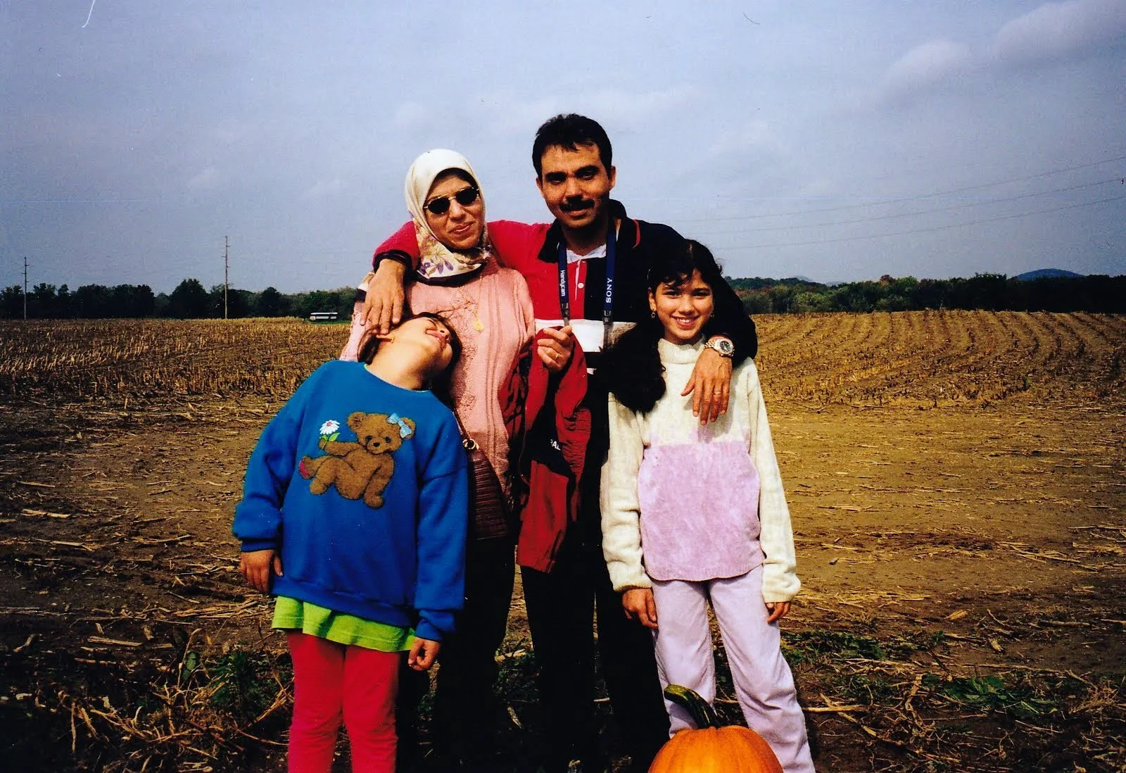 A family of five standing outdoors on a farm field with pumpkins in autumn. The woman is wearing sunglasses and a headscarf. The man has a camera around his neck. Two young girls are in front, one wearing a blue sweater with a teddy bear and the other wearing a light-colored sweater with a pink apron. All are smiling and enjoying the weather.