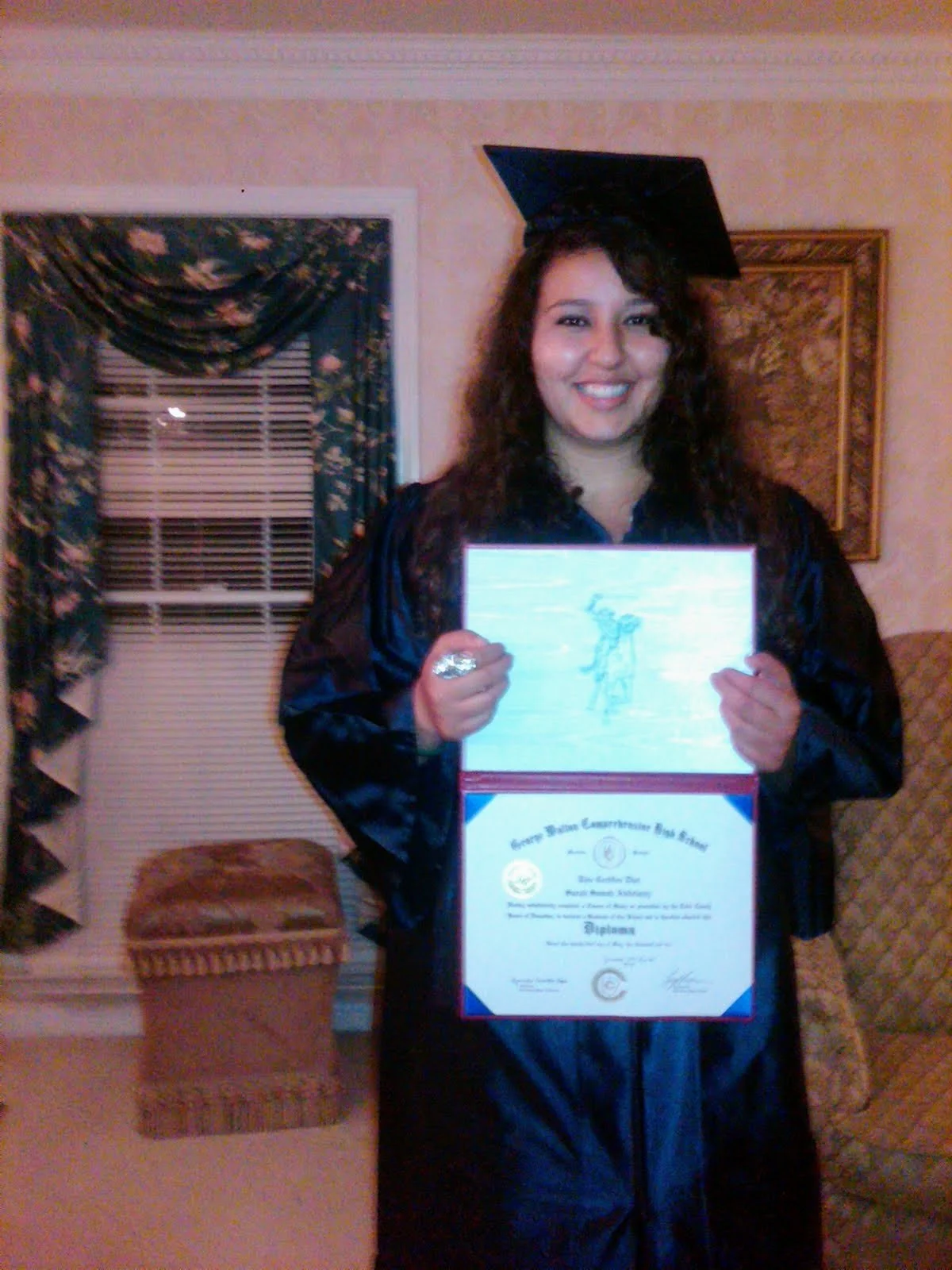 Young woman in graduation cap and gown holding a diploma and certificate, standing in a decorated room.