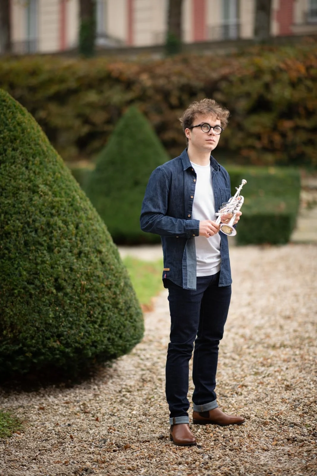 Jeune homme à lunettes tenant une trompette piccolo à l'extérieur parmi des buissons verts et un sol de gravier
