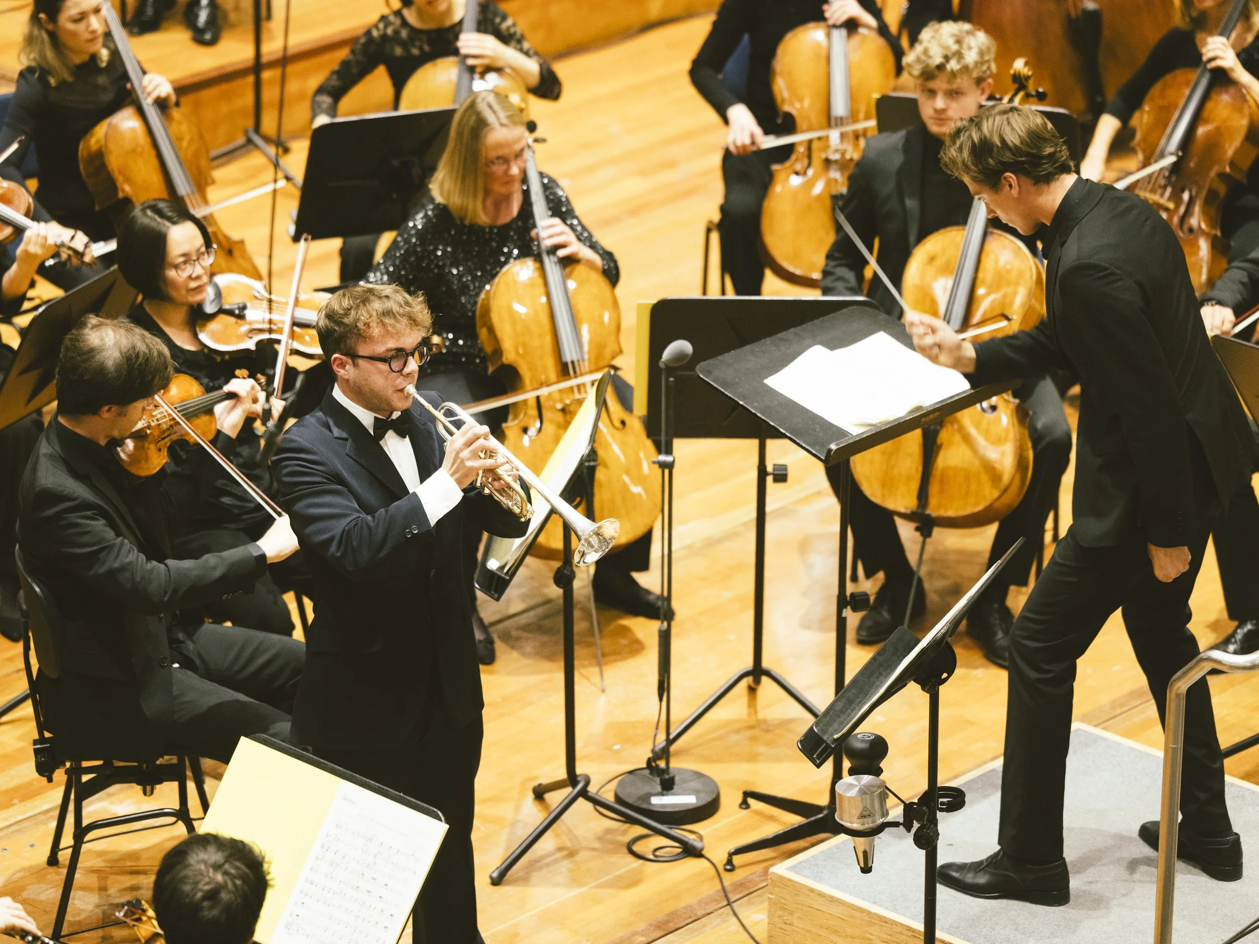 Un groupe de musiciens jouant dans un orchestre symphonique, dirigé par un chef d'orchestre. Divers instruments à cordes et un trompettiste soliste sont visibles, dans une salle de concert 
