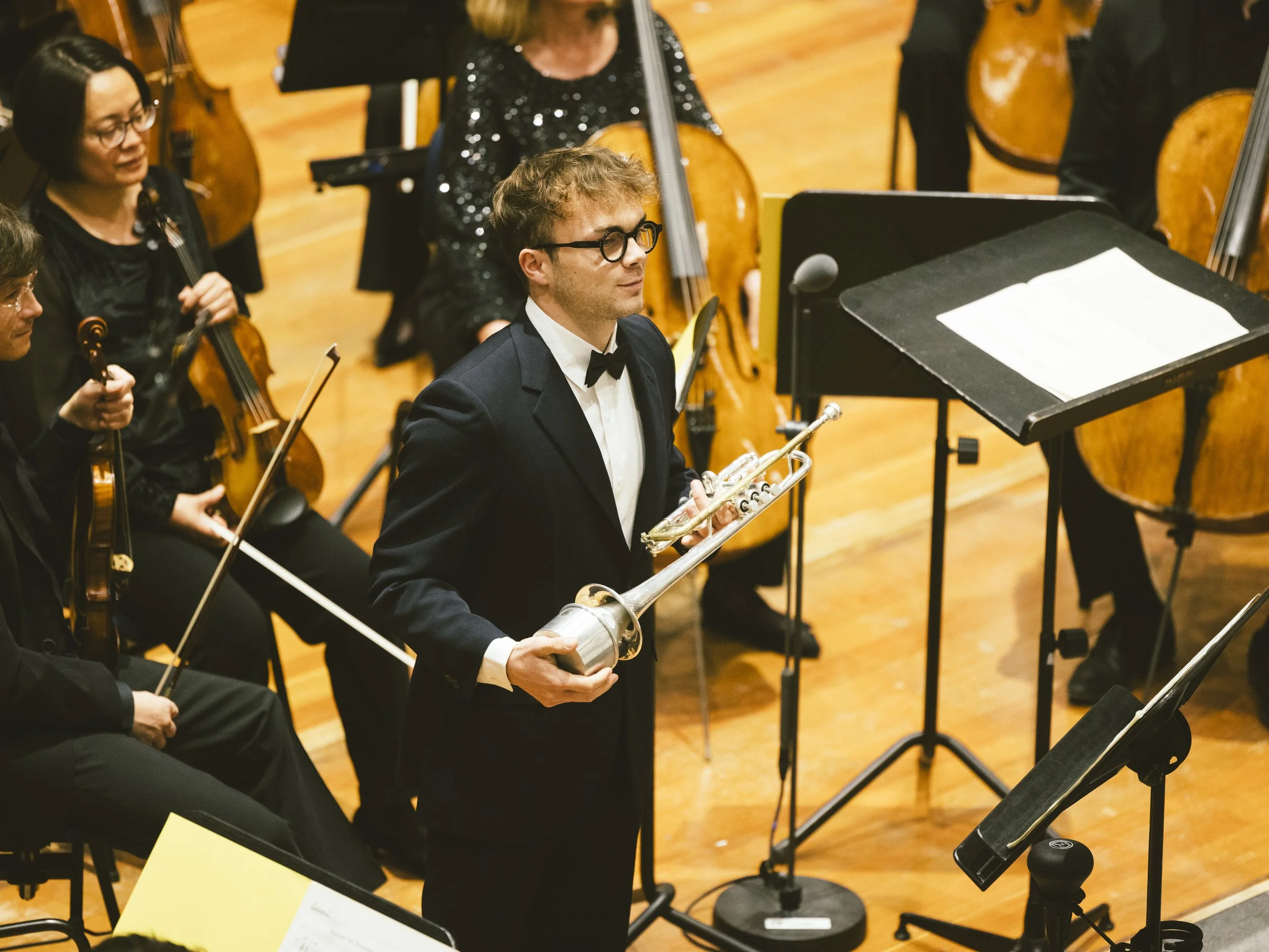 Un jeune homme en smoking tient une trompette lors d'un concert d'orchestre, entouré d'autres musiciens 