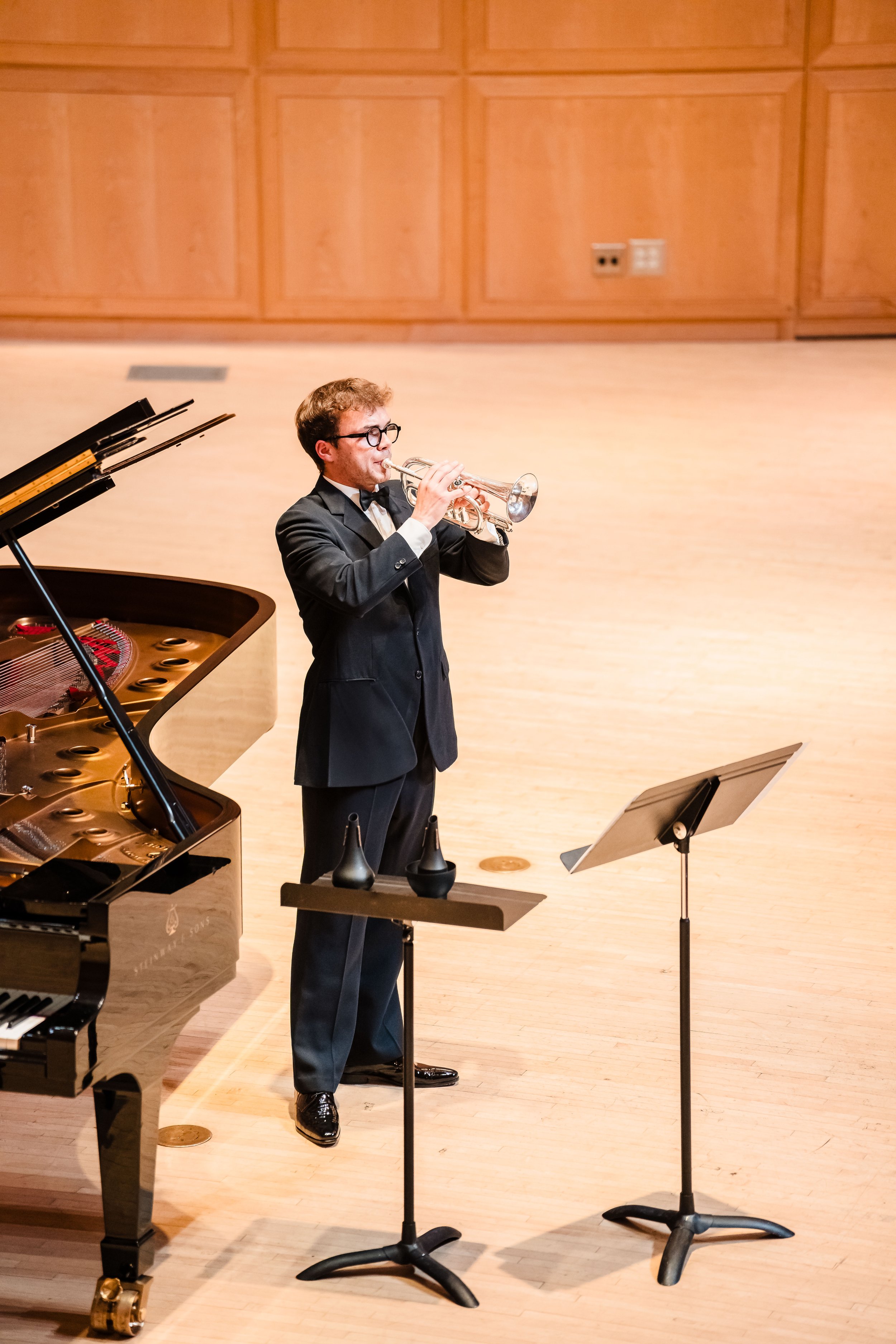 Un musicien en costume noir joue de la trompette dans une salle de concert avec un piano à queue à gauche et un pupitre à partition devant lui.