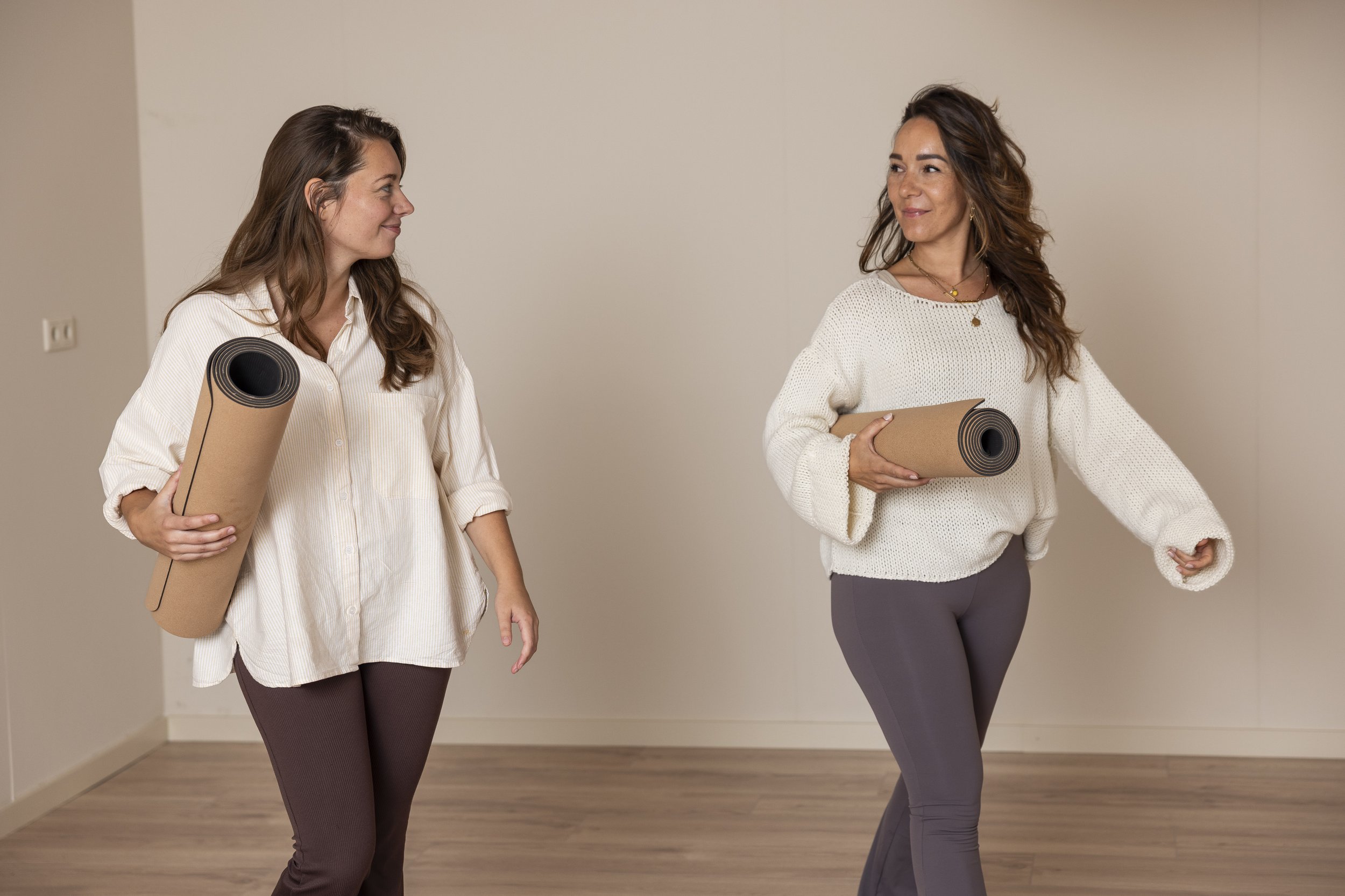 Twee vrouwen lopen samen binnenshuis, glimlachend naar elkaar, elk met een opgerolde yogamat in comfortabele yogakleding, bij Roots Yoga in Deurne.