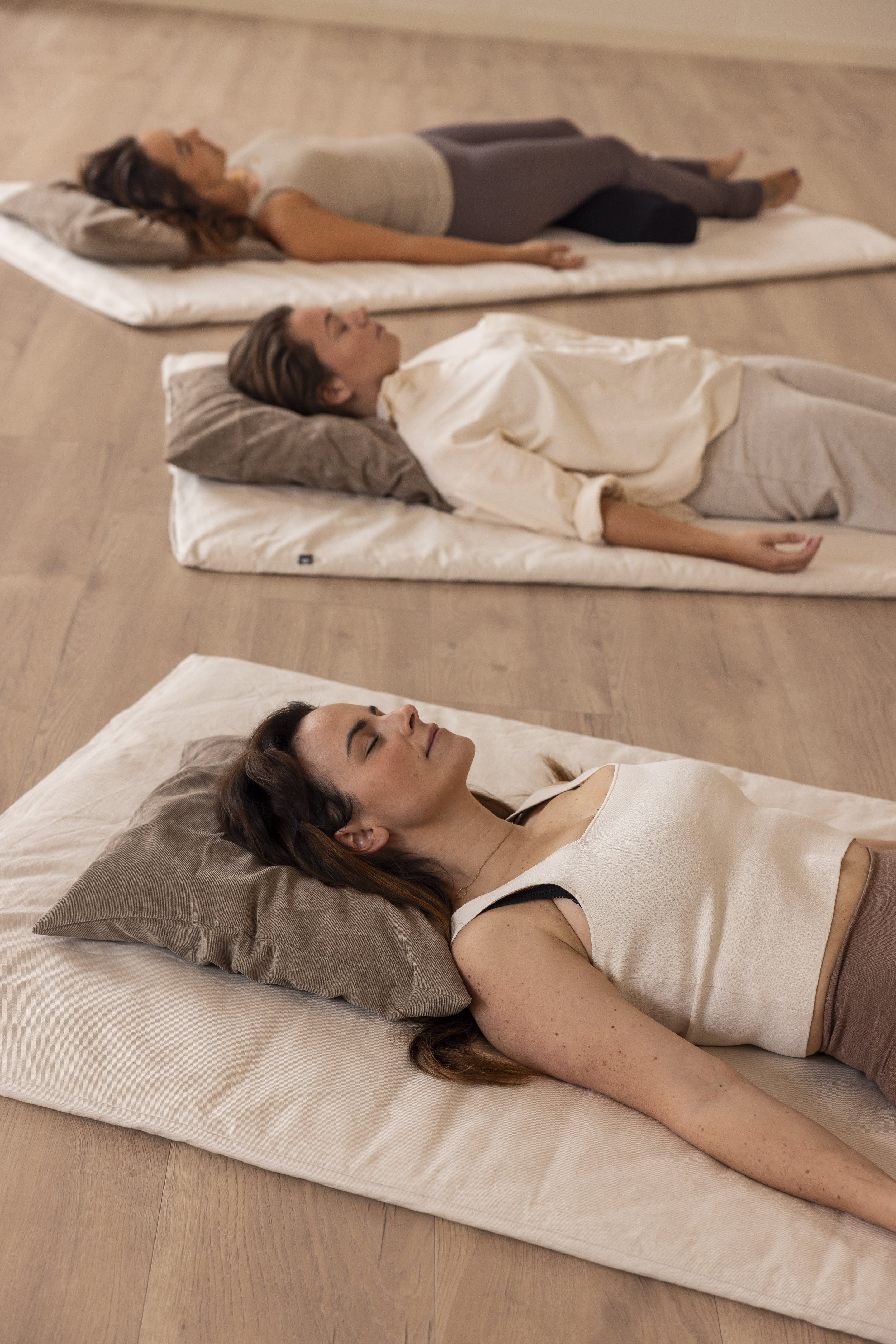 Drie vrouwen liggend op yogamatten tijdens een meditatie- of ontspanningssessie, met gesloten ogen in een ruime studio met houten vloer bij Roots Yoga in Deurne.