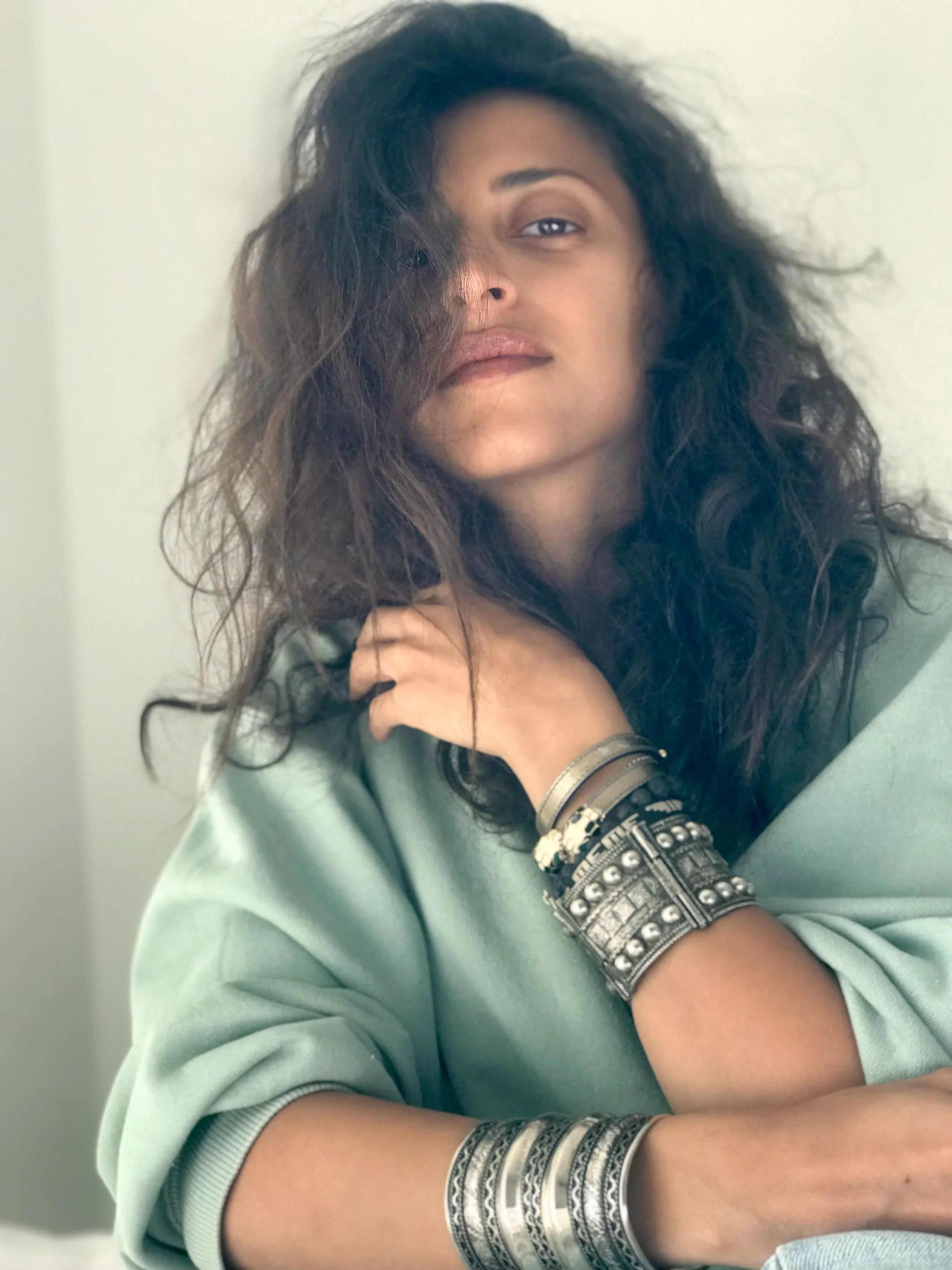 A woman with curly dark hair and light skin poses with her arm resting on her shoulder, wearing multiple silver bracelets and bangles, dressed in a light green top, with a neutral expression, against a plain background.