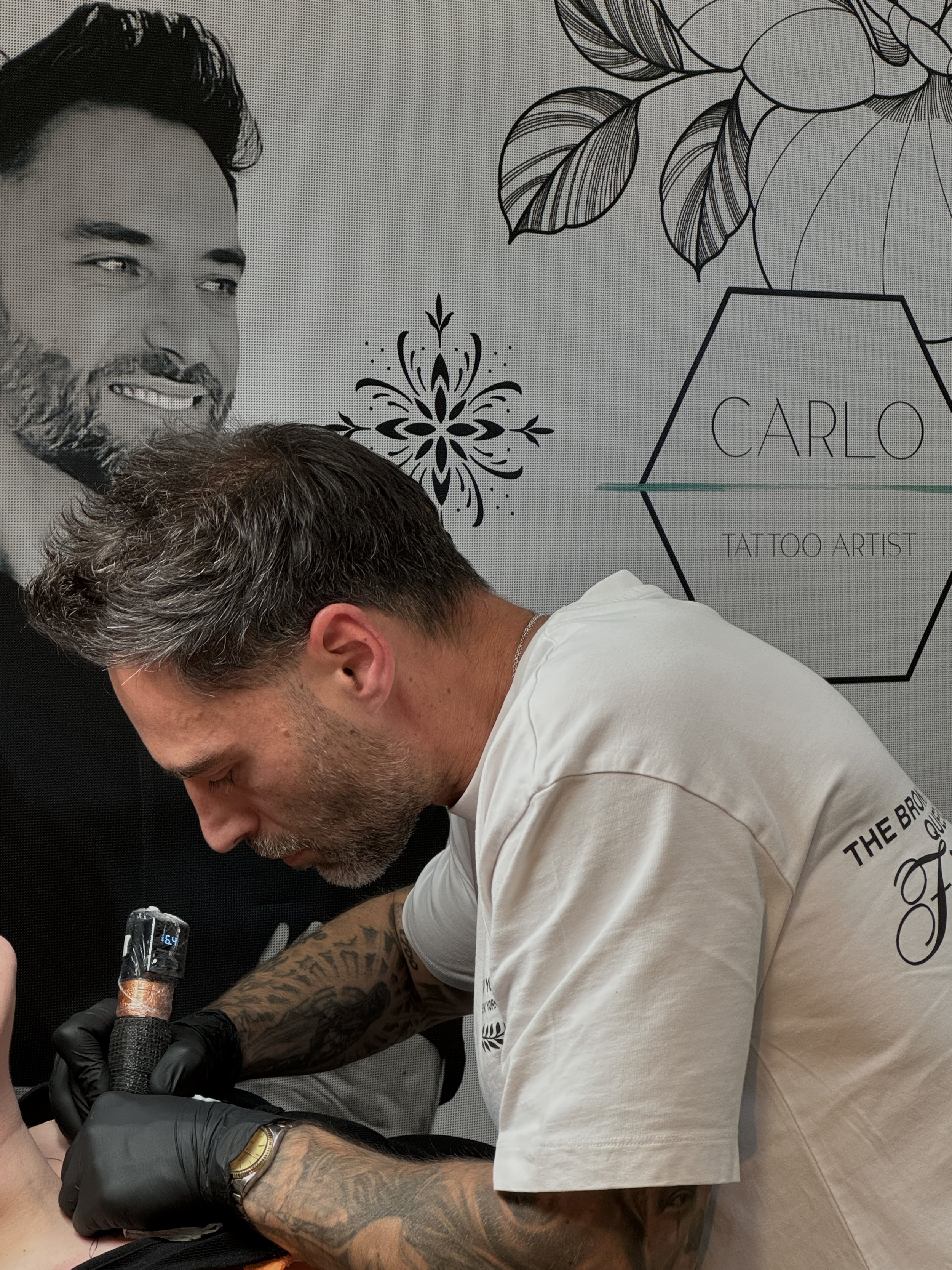 Carlo Tattoo artist with tattoos on his arm, wearing a white T-shirt and black gloves, working on a person's tattoo.