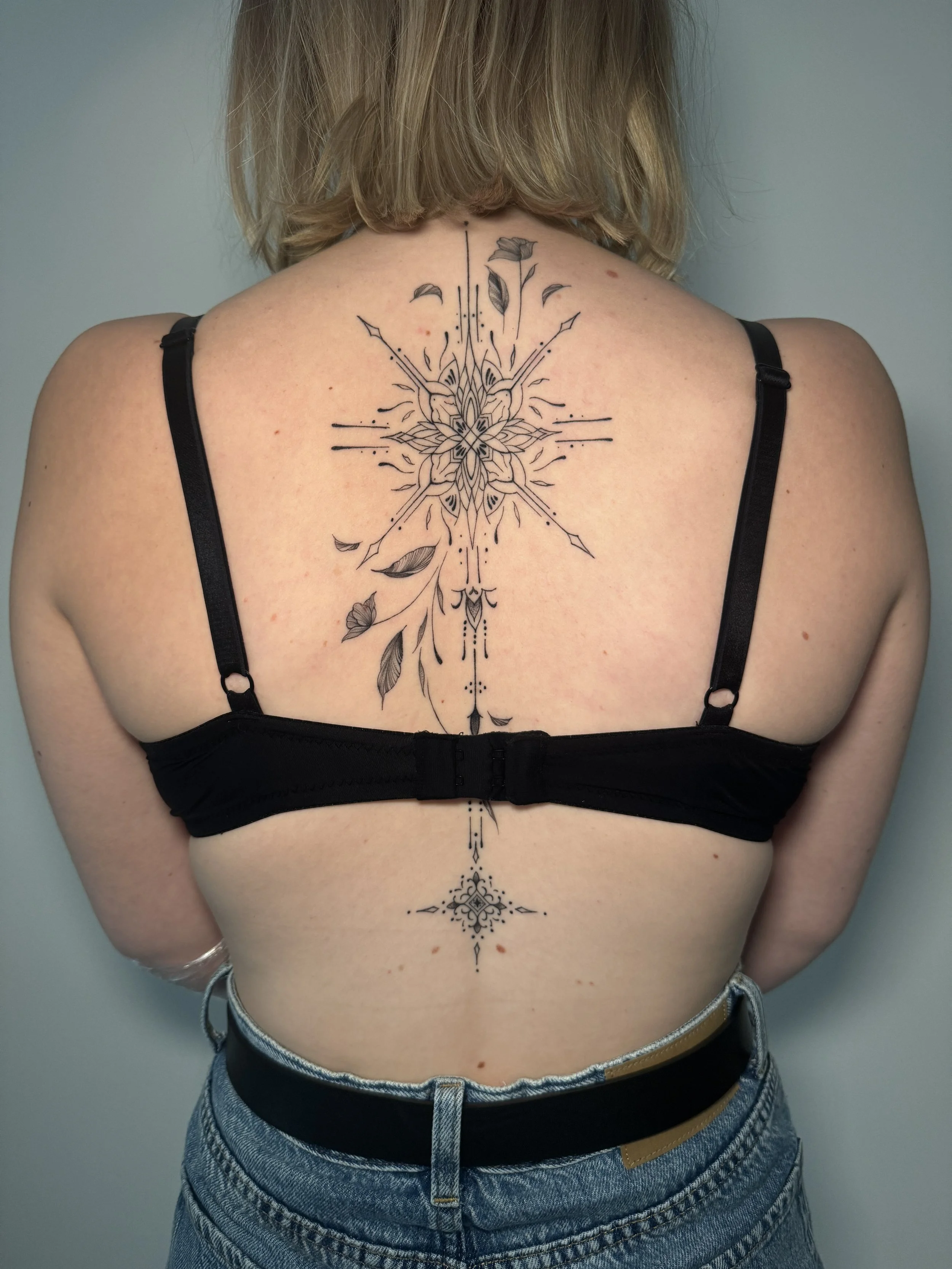 Back view of a woman with a large geometric tattoo featuring floral elements and lines, extending from her upper back to her lower back.