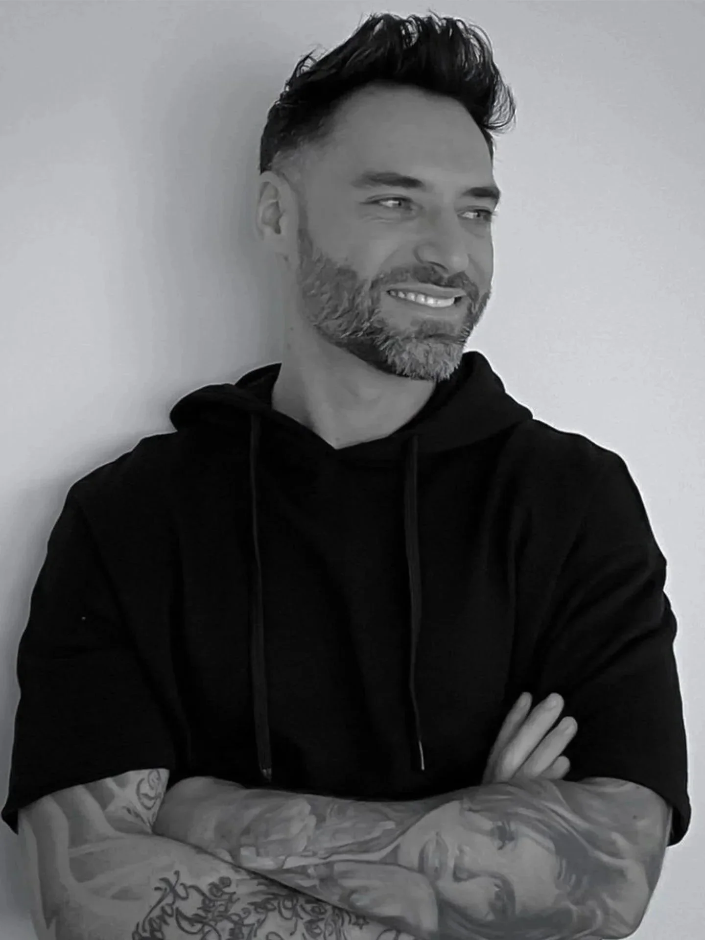 Black and white photo of a man with styled hair and a beard, smiling and looking to the side, with his arms crossed and tattoos on his forearms, wearing a black hoodie.