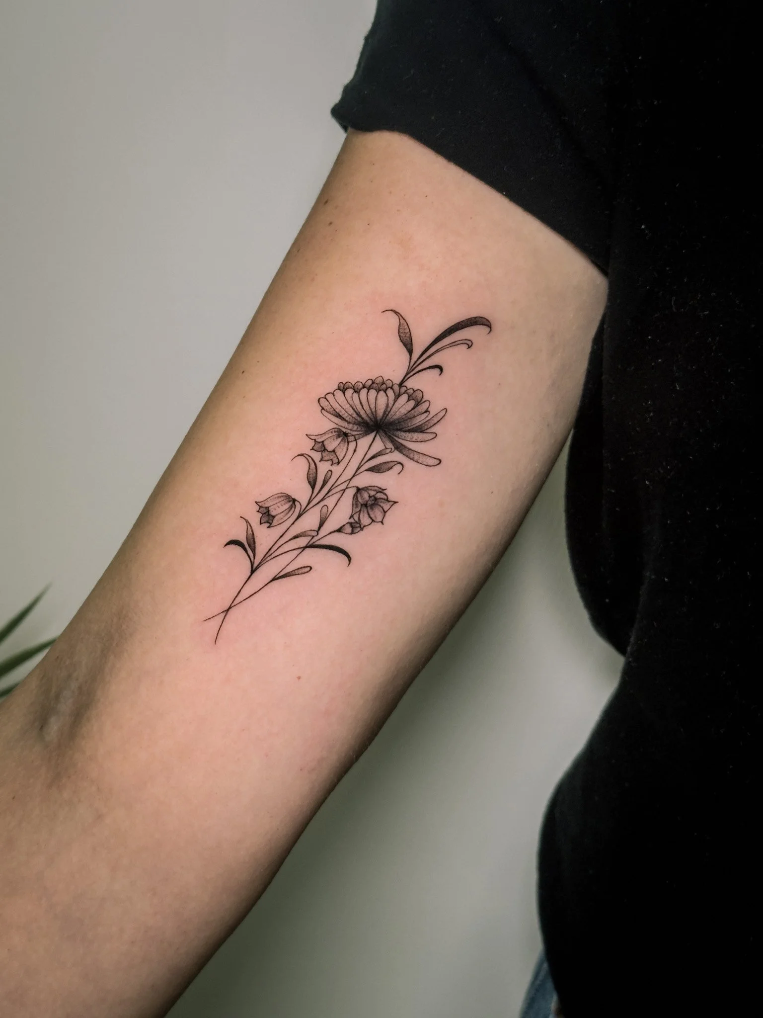 A black and grey floral tattoo on a person's forearm featuring a large daisy-like flower with smaller surrounding flowers and leaves.