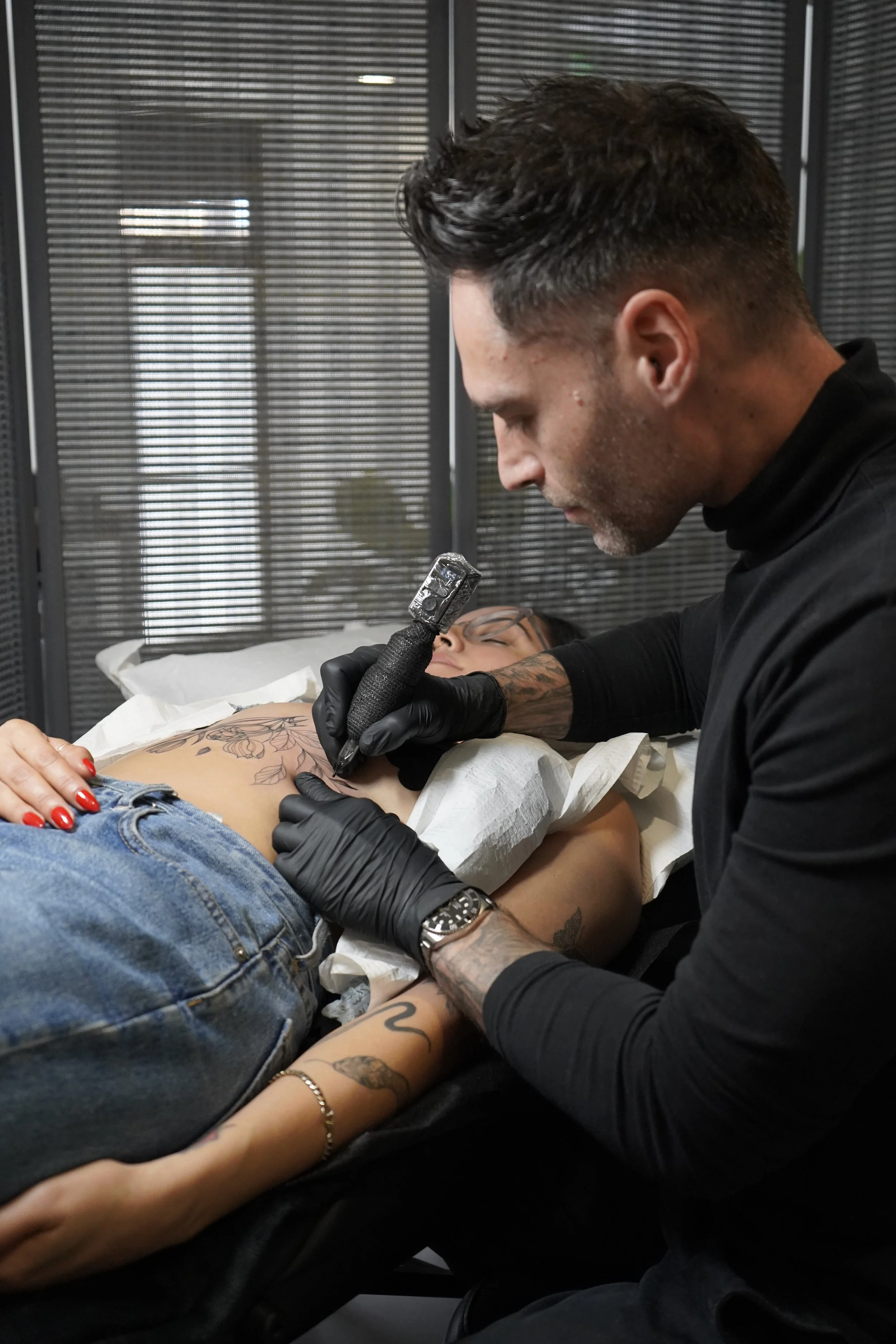 Carlo tattoo artist creating a floral tattoo on a woman lying on a bed, using a tattoo machine in a professional studio.