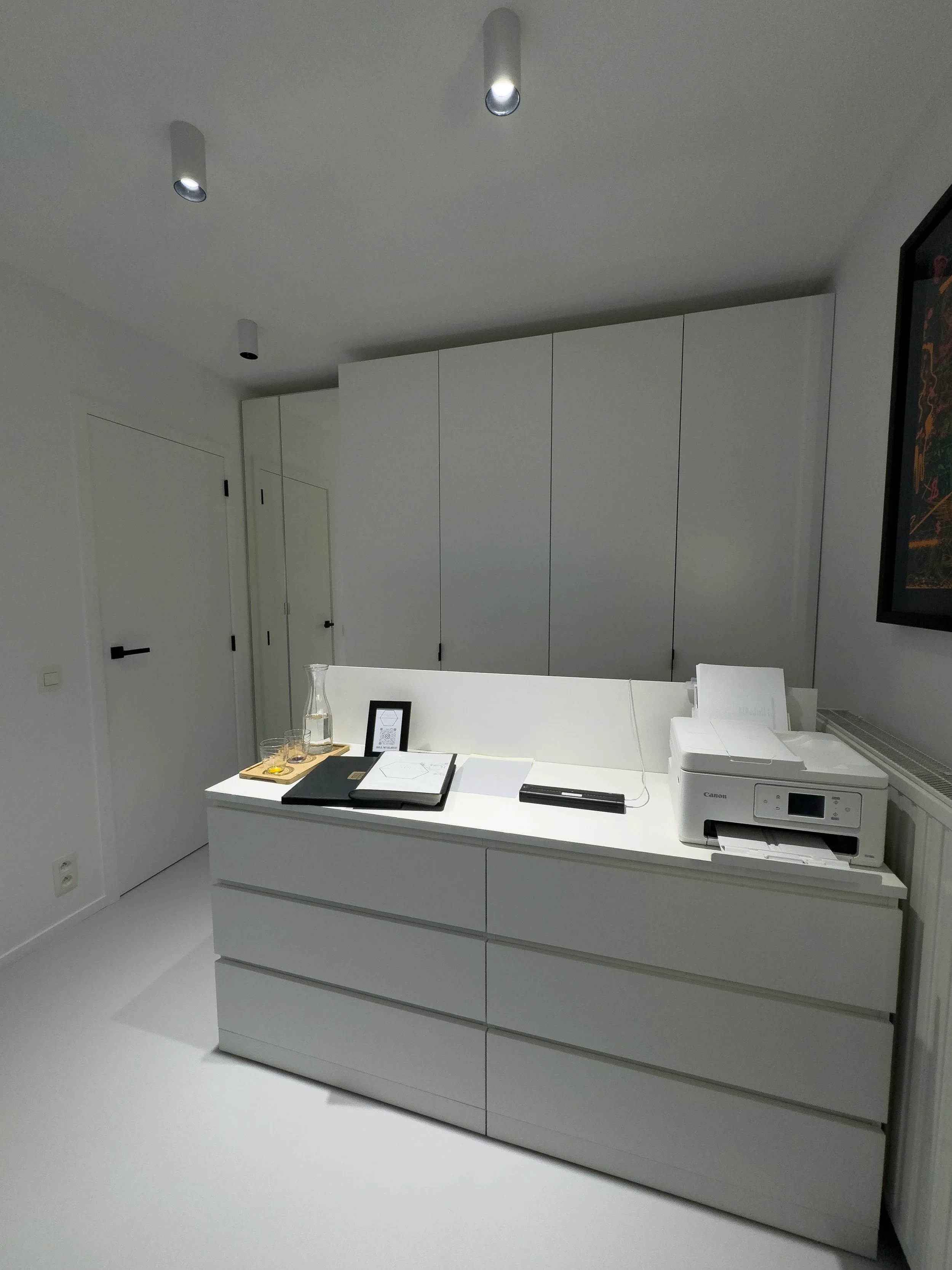 Modern tattoo space with a white countertop, printer, documents, water pitcher, and glasses, with cabinets and a wall-mounted tv.