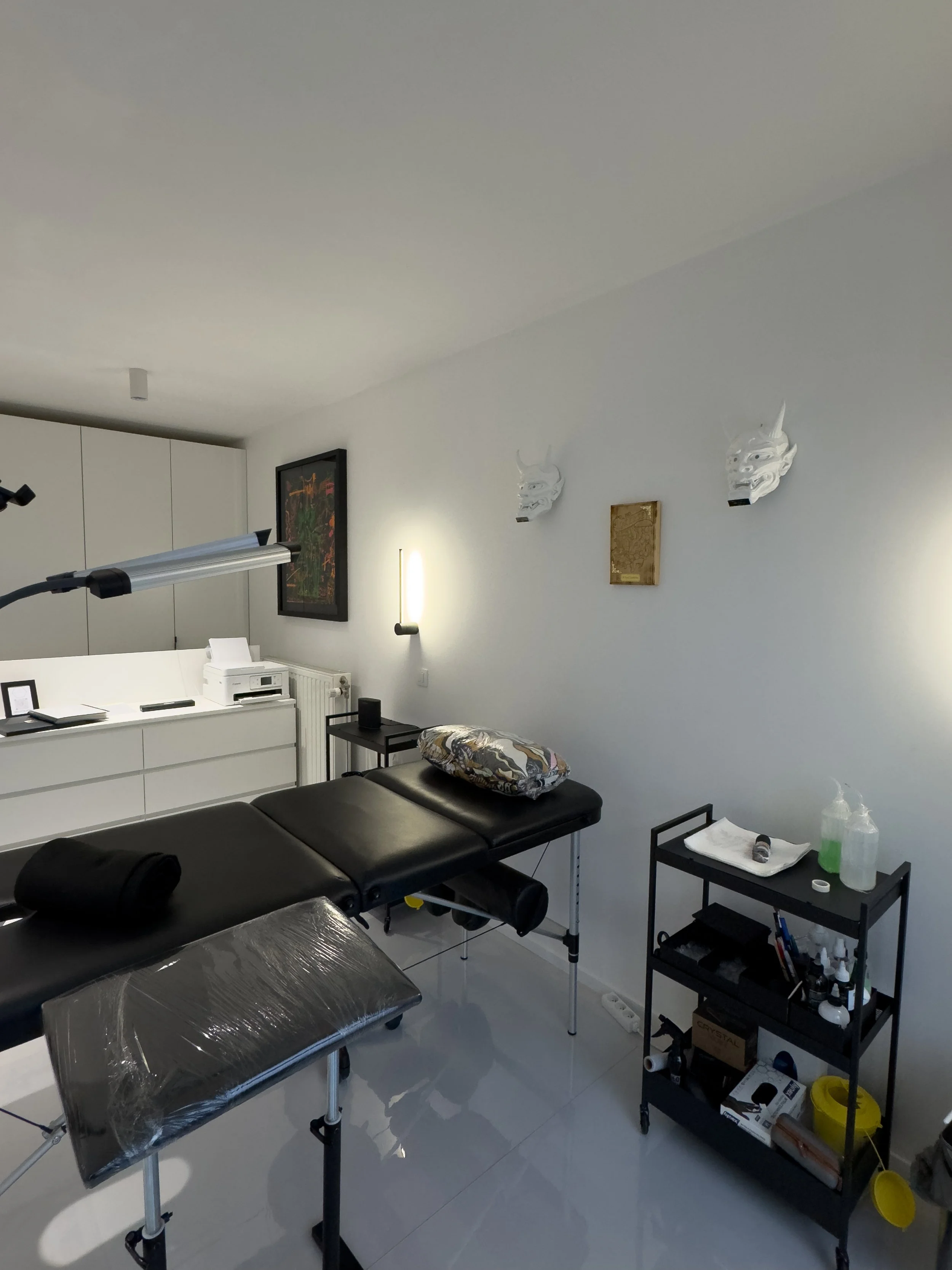 Tattoo studio with a black massage table, tattoo supplies on a shelf, and wall decorations including two white masks and a framed artwork.
