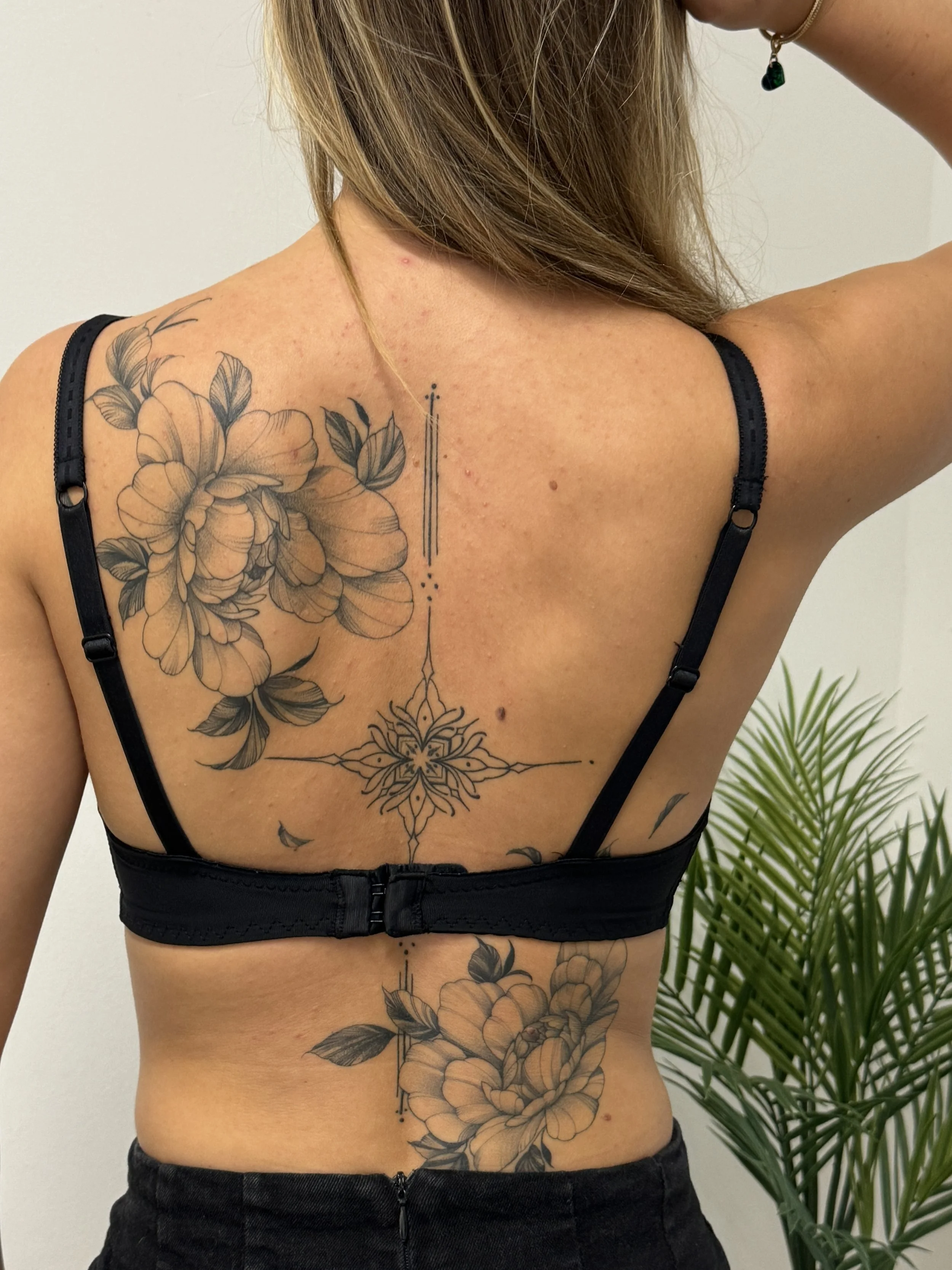 A woman with a back tattoo of large peony floral designs, geometric lines, and dots, wearing a black bra.