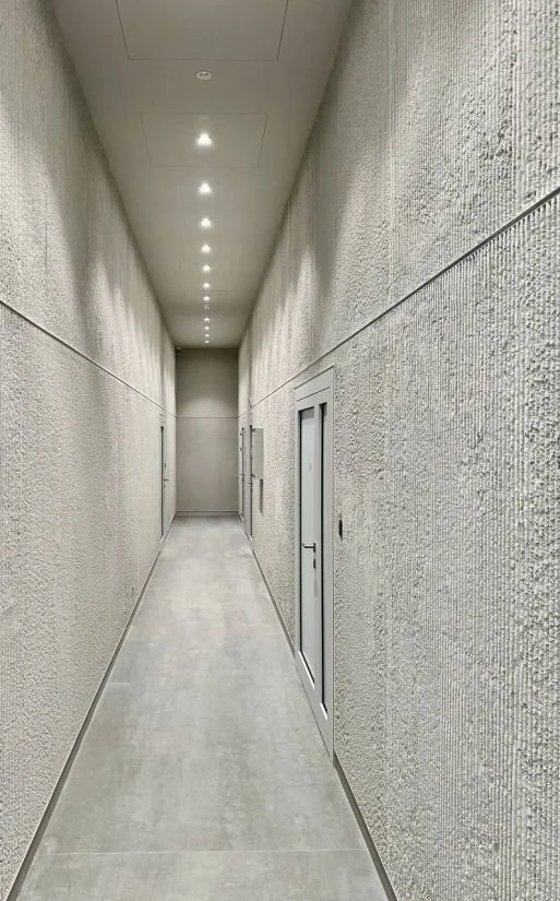 Empty, narrow corridor with textured plaster white walls, a series of ceiling lights, and two gray doors on the right side.
