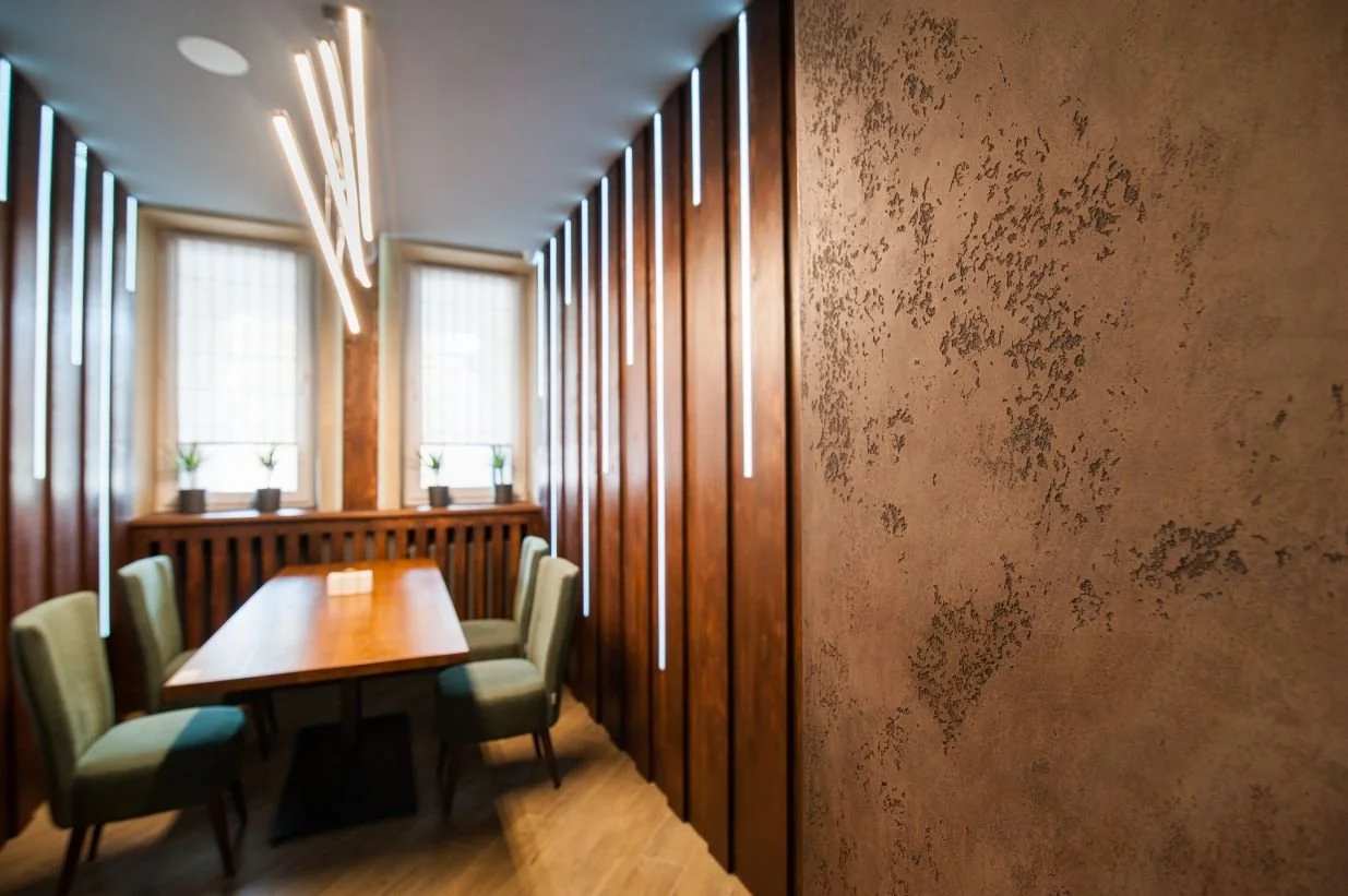 A modern dining room with a wooden table, six green chairs, three windows with small potted plants, vertical wooden panels, and unique vertical light fixtures on the ceiling.