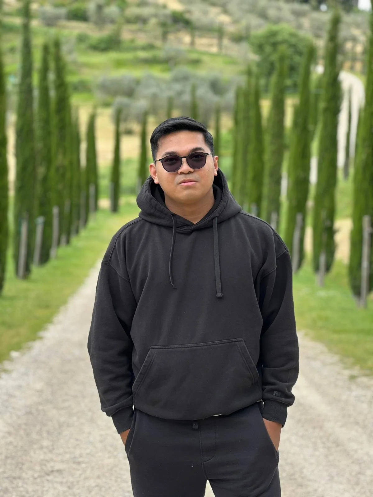 A young man wearing sunglasses, a black hoodie, and black pants stands on a gravel path surrounded by rows of green, moss-covered trees in a park or garden.