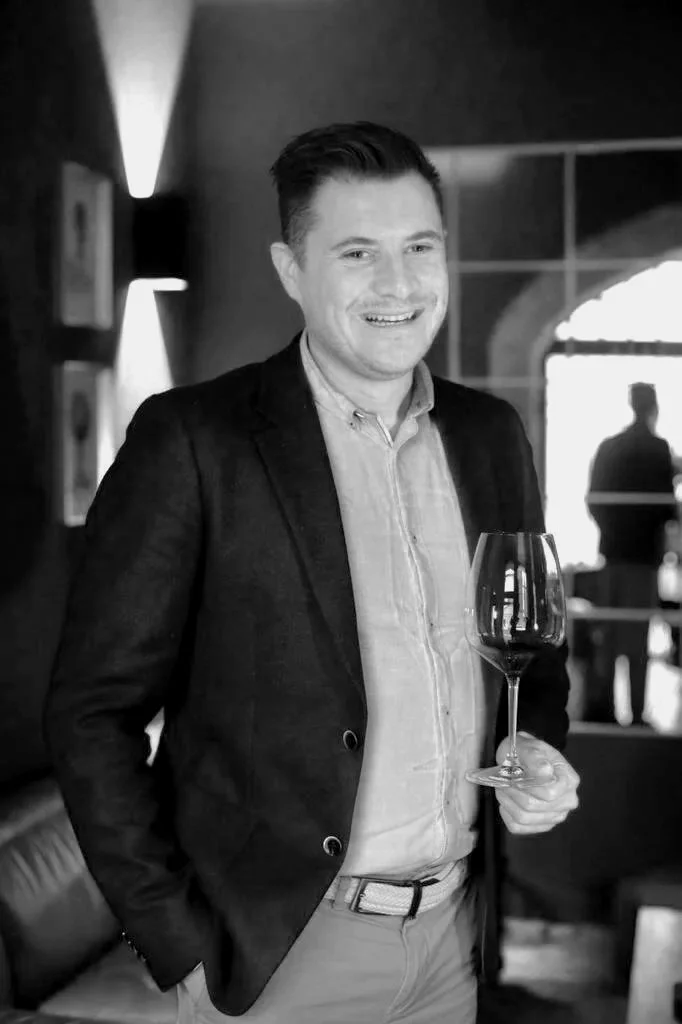 A man in a blazer and dress shirt holding a wine glass in a restaurant or bar setting, smiling.