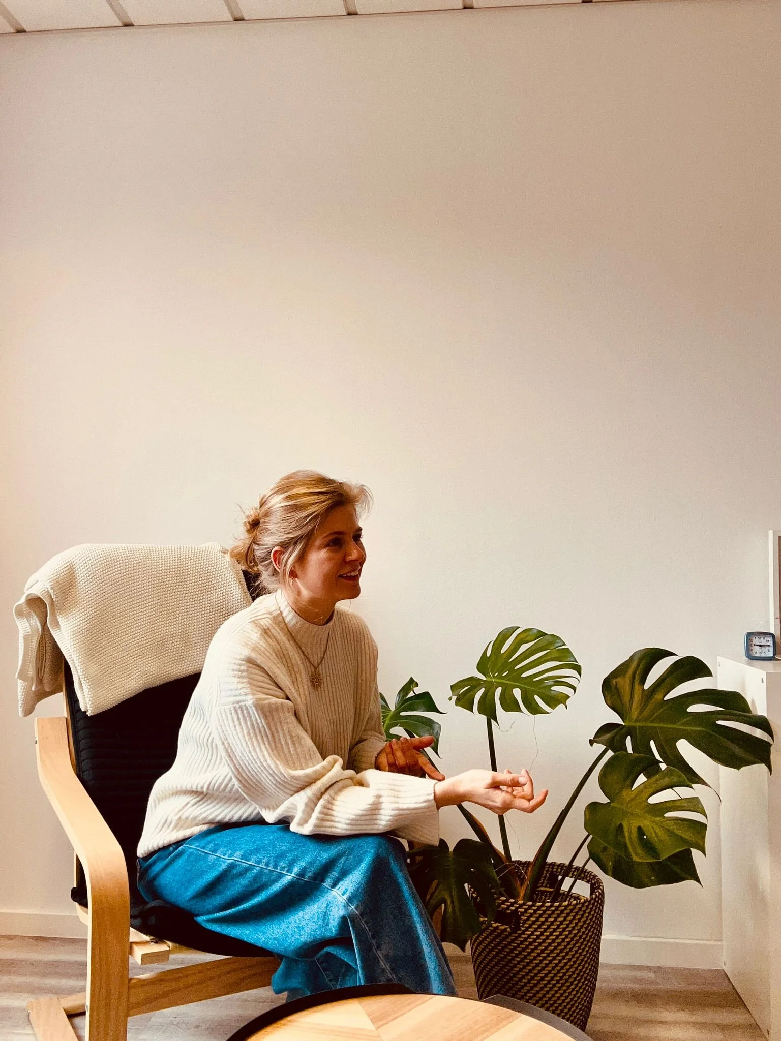 Eine Frau sitzt in einem Raum auf einem Stuhl, trägt einen beigen Pullover und blaue Jeans, neben ihr befinden sich große Monstera-Pflanzen in einem Korb.