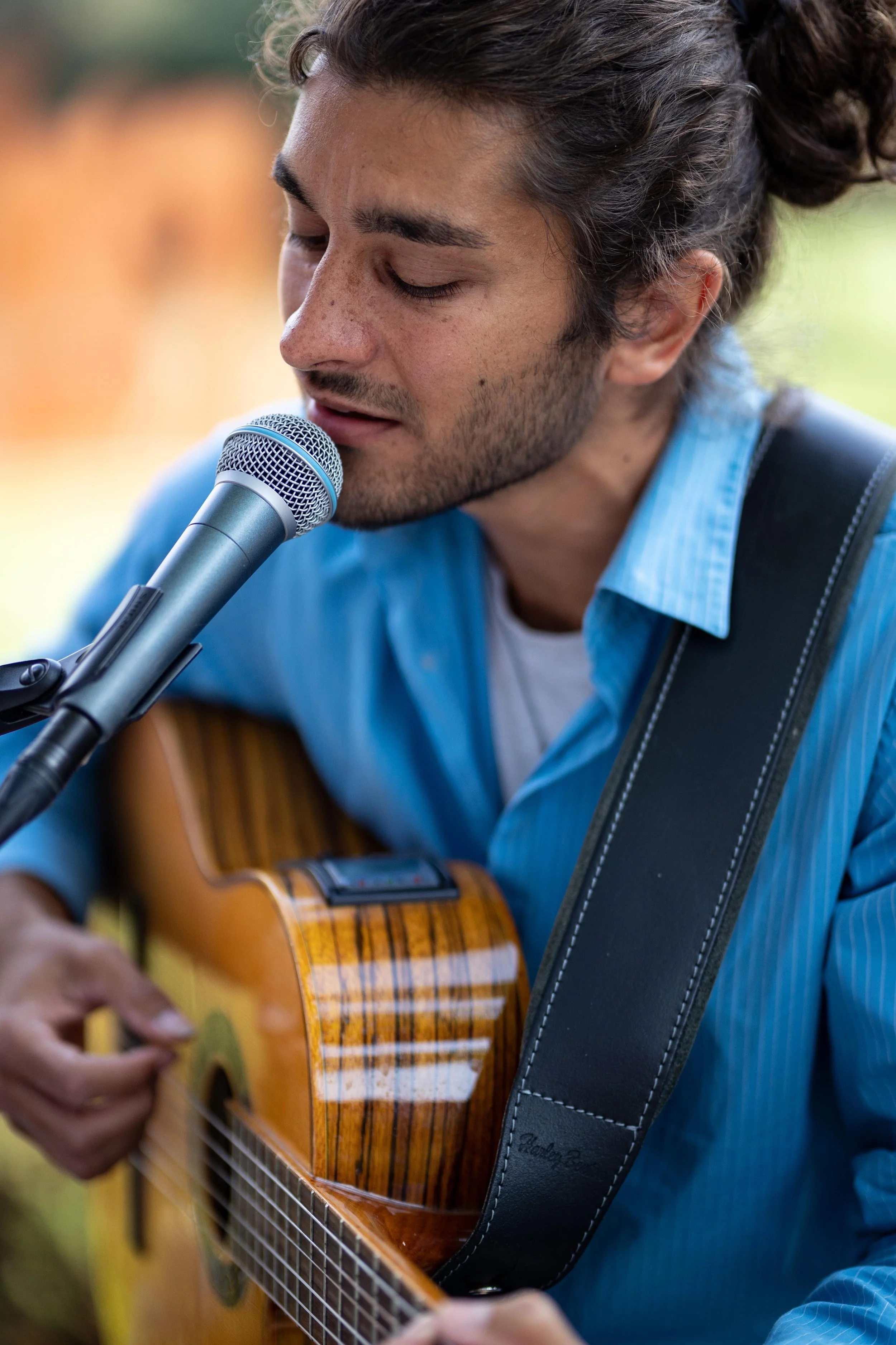 Ein junger Mann mit dunklem, gewelltem Haar und Bart spielt Gitarre und singt in ein Mikrofon, während er draußen im Freien unter einem Himmel mit natürlichem Licht sitzt.