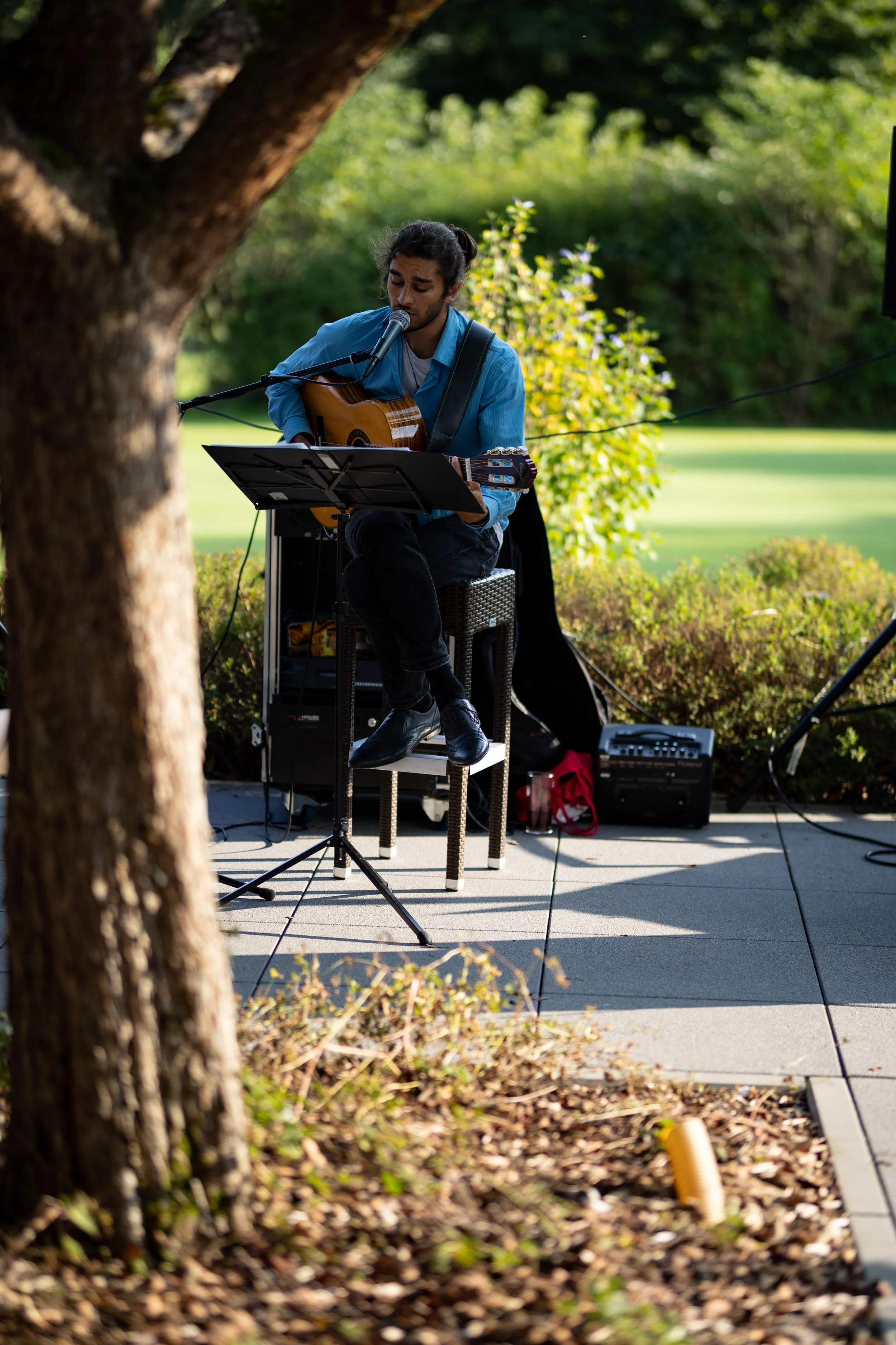 Ein Mann spielt akustische Gitarre und singt in einem Outdoor-Garten, mit Bäumen und grünem Rasen im Hintergrund.