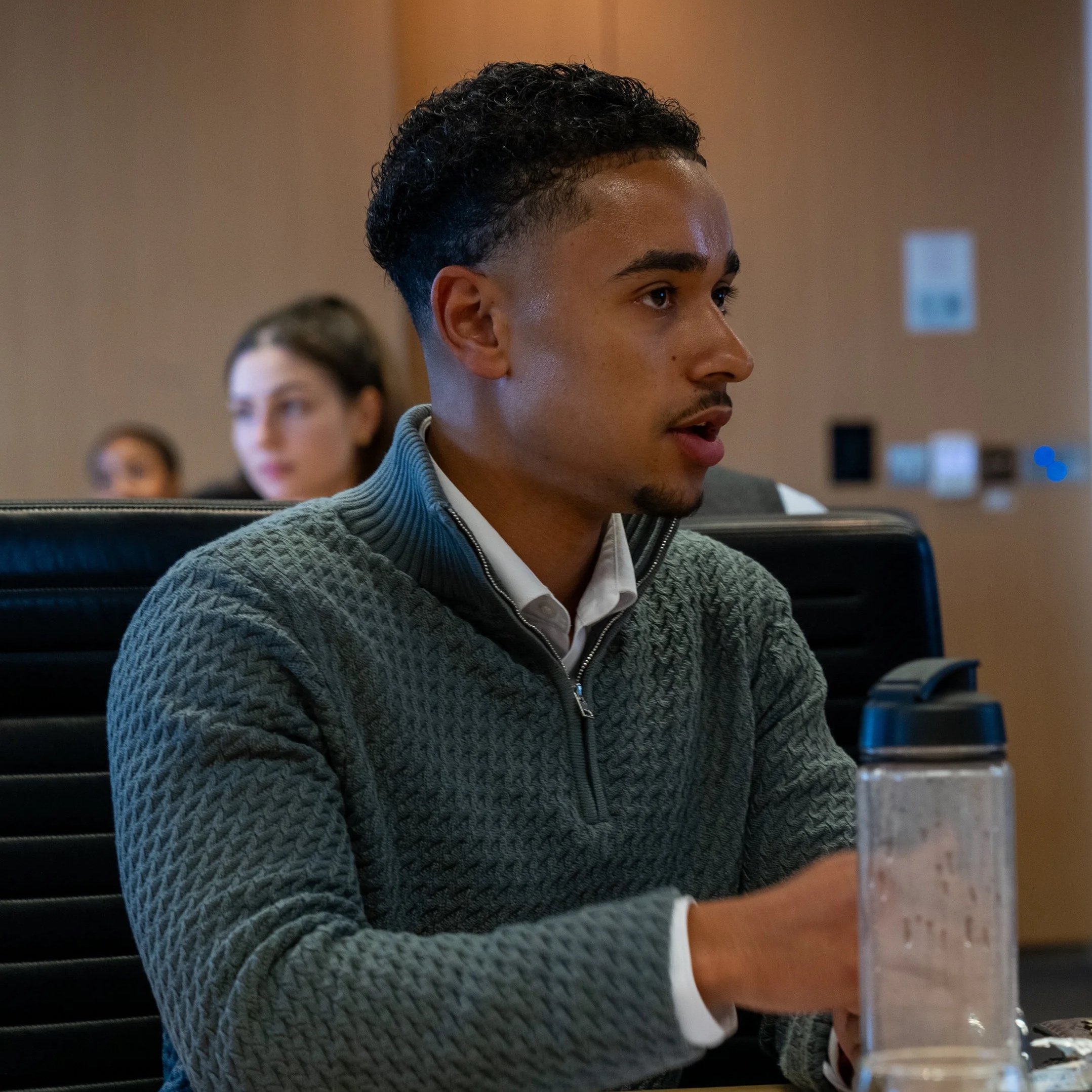 Young man sitting at a conference table with a water bottle in front of him, wearing a grey sweater and white shirt, focused on a presentation or speaker.