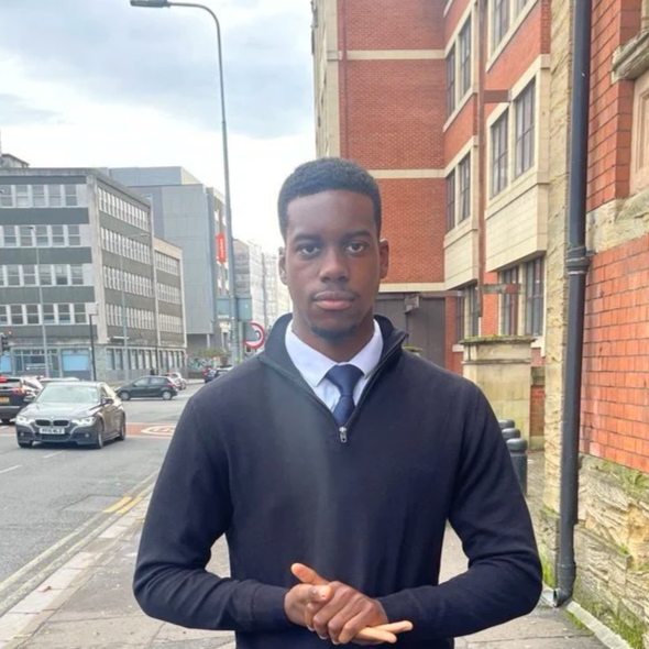 A young man in formal attire standing on a city sidewalk with buildings and parked cars in the background.