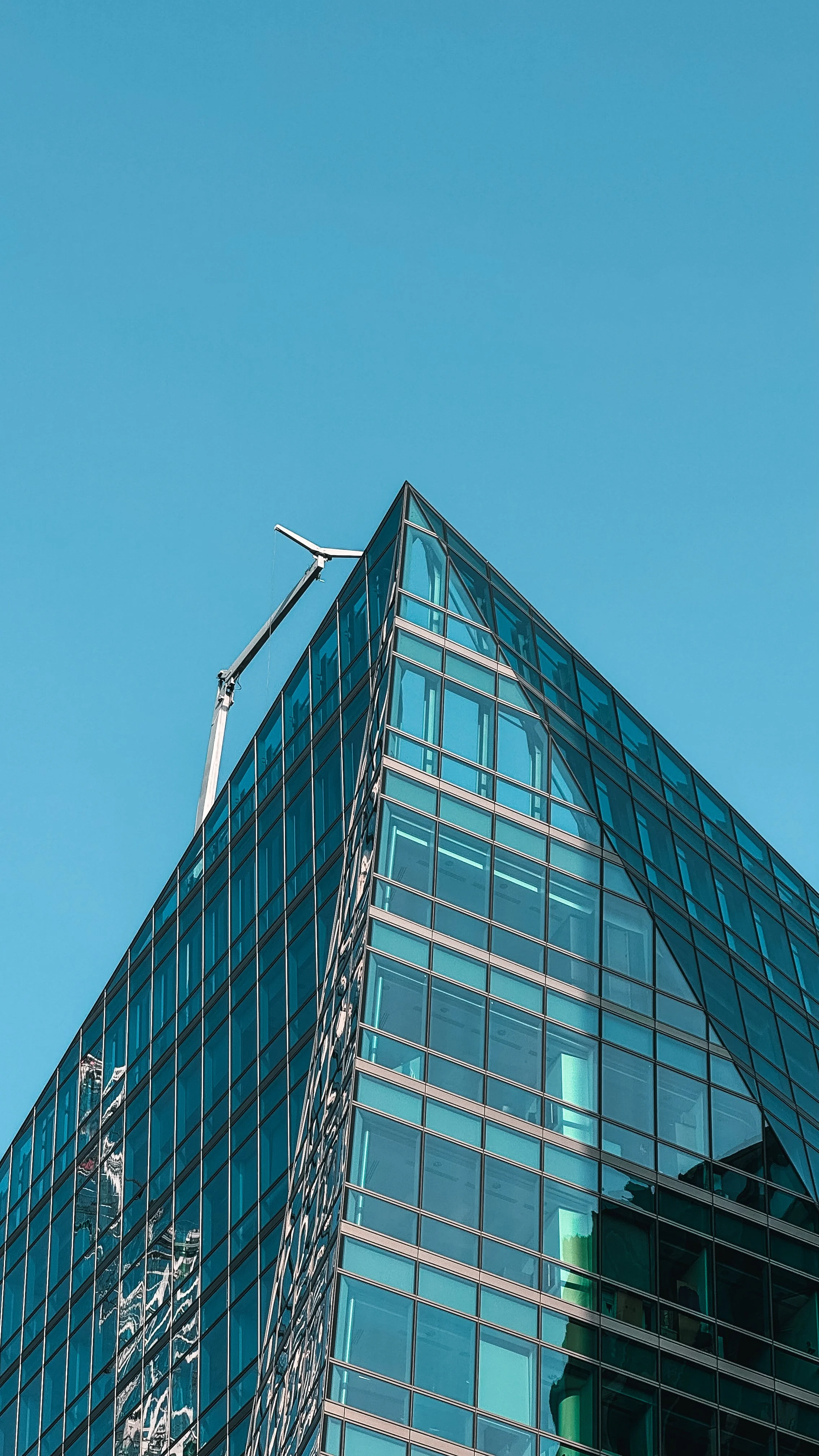 Modern glass skyscraper with a crane on top against a clear blue sky.