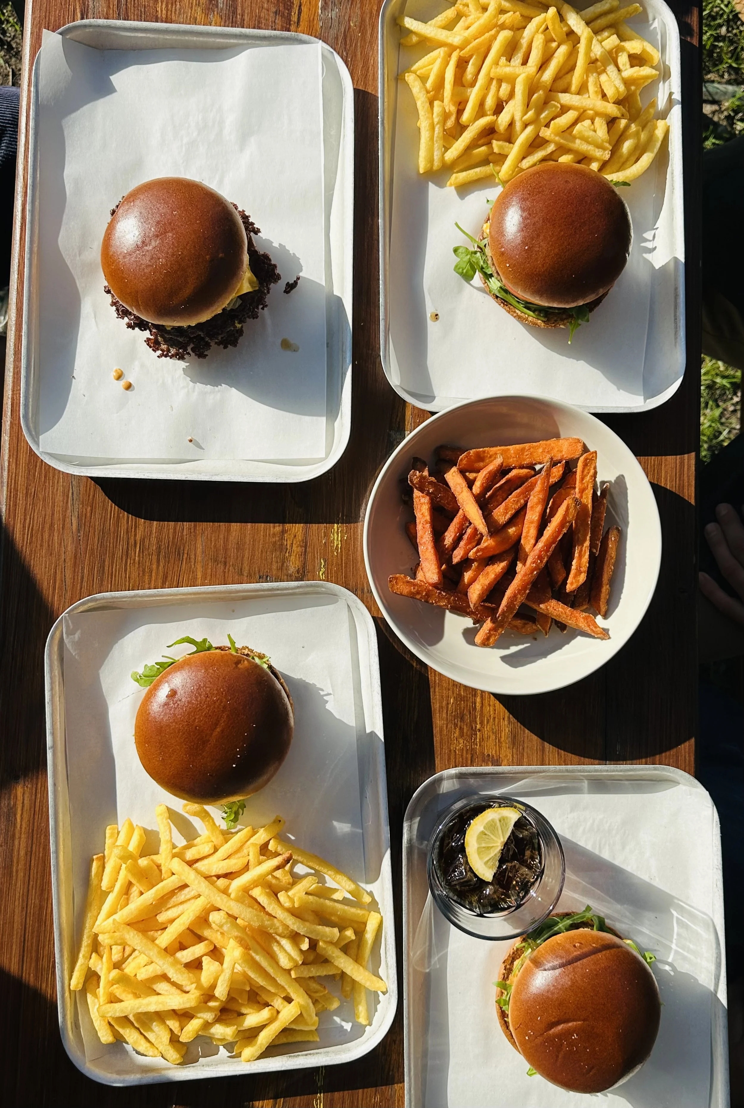Verschiedene Burger mit Pommes, Süßkartoffelpommes und einem Getränk mit Zitronenscheibe auf einem Holztisch.