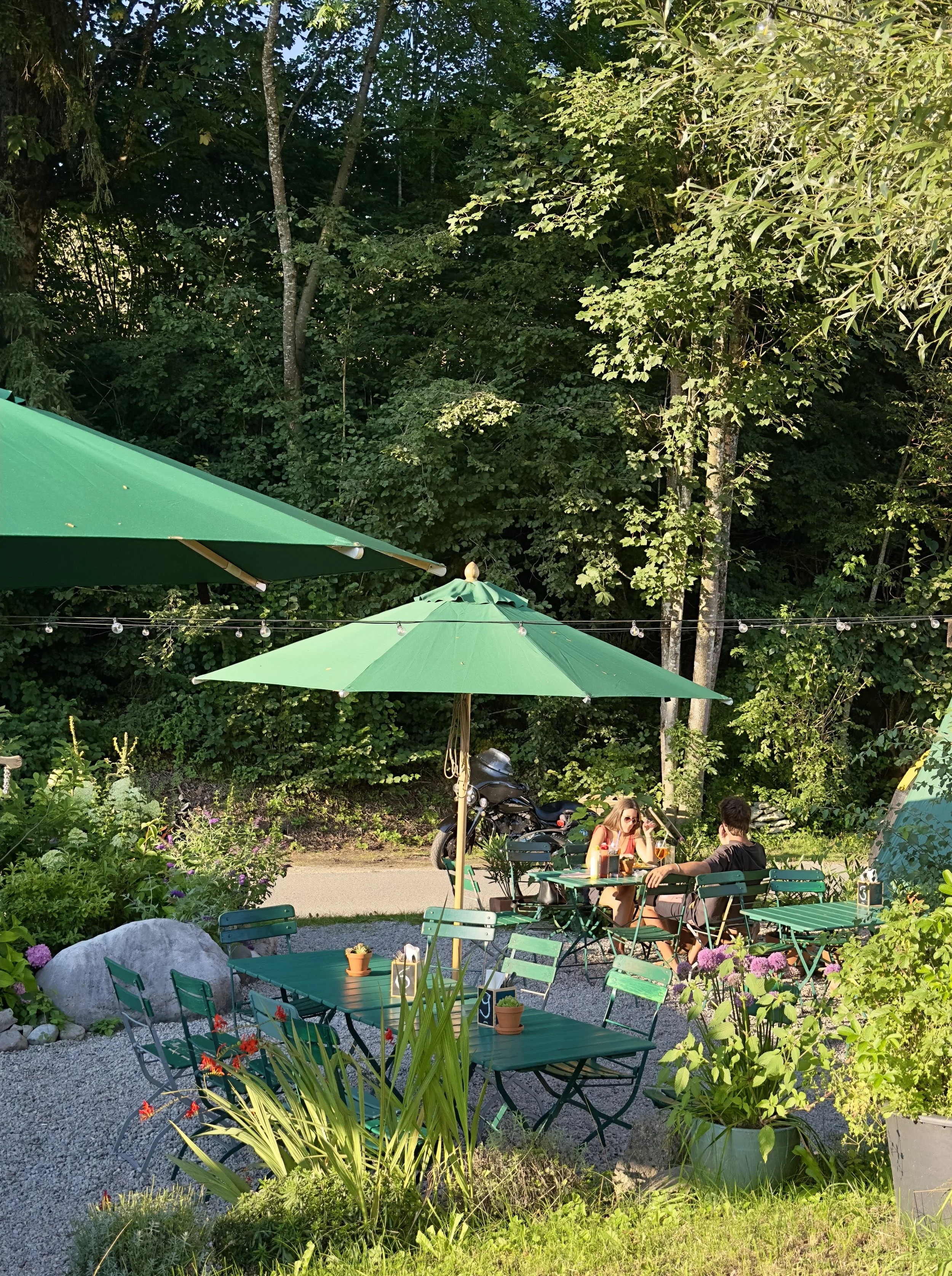 Ein Garten oder eine Terrasse mit mehreren grünen Tischen und Stühlen, einige mit Sonnenschirmen. Zwei Menschen sitzen an einem Tisch, trinken und sprechen. Es gibt einige Pflanzen und große Steine, im Hintergrund Bäume, und es hängt eine Lichterkette.