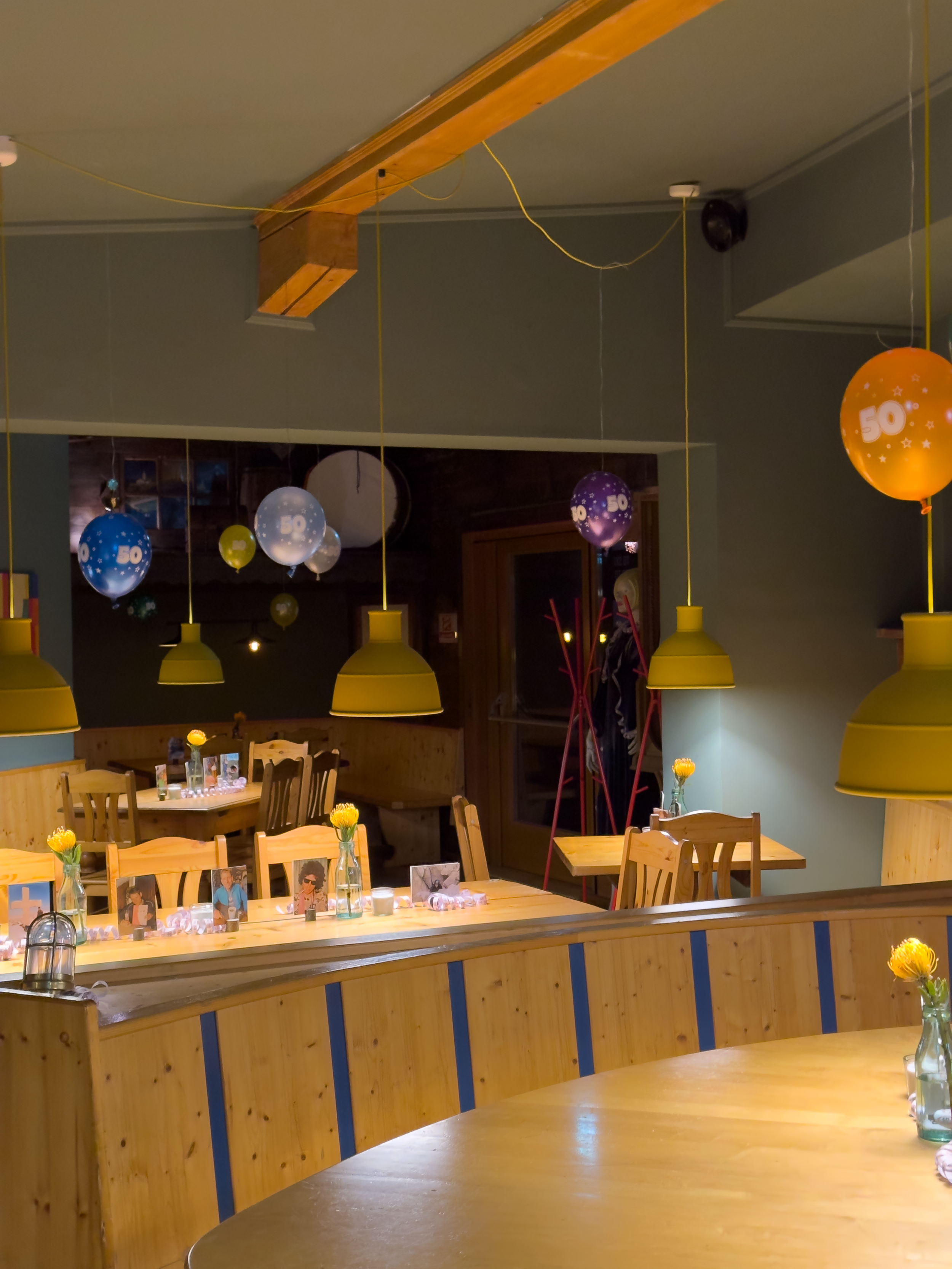 Innenansicht eines Restaurants mit Holztischen und Stühlen. Deckenleuchten in Gelb, bunte Luftballons mit der Zahl 50 hängen an der Decke. Auf den Tischen sind Vasen mit gelben Blumen und Fotos.
